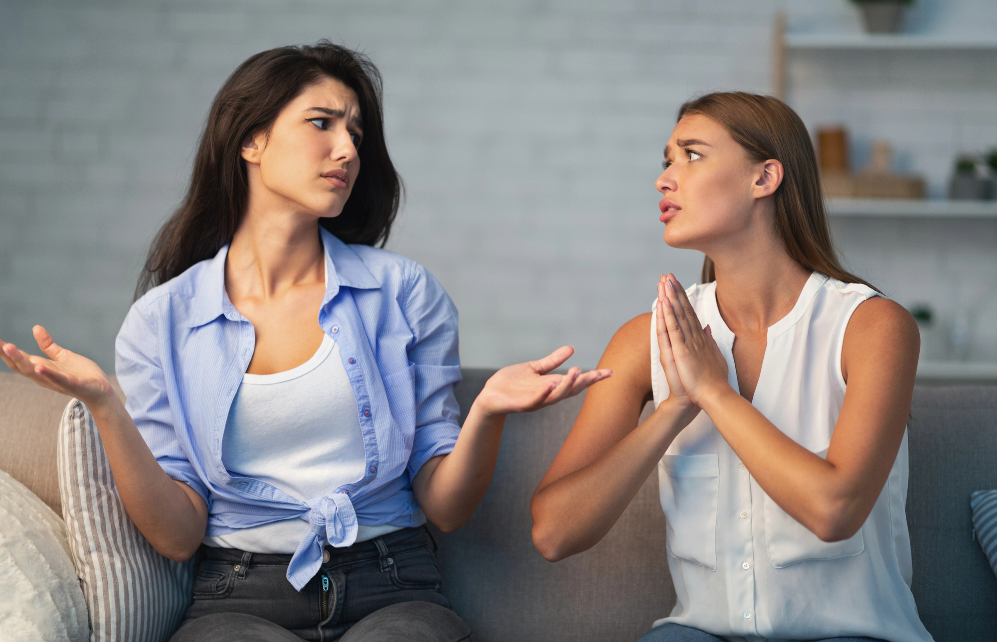 Two women are sitting on a couch. The woman on the left is frowning with her arms out. The woman on the right has her hands together and is looking at the other woman with a pleading expression. They appear to be having a serious conversation.