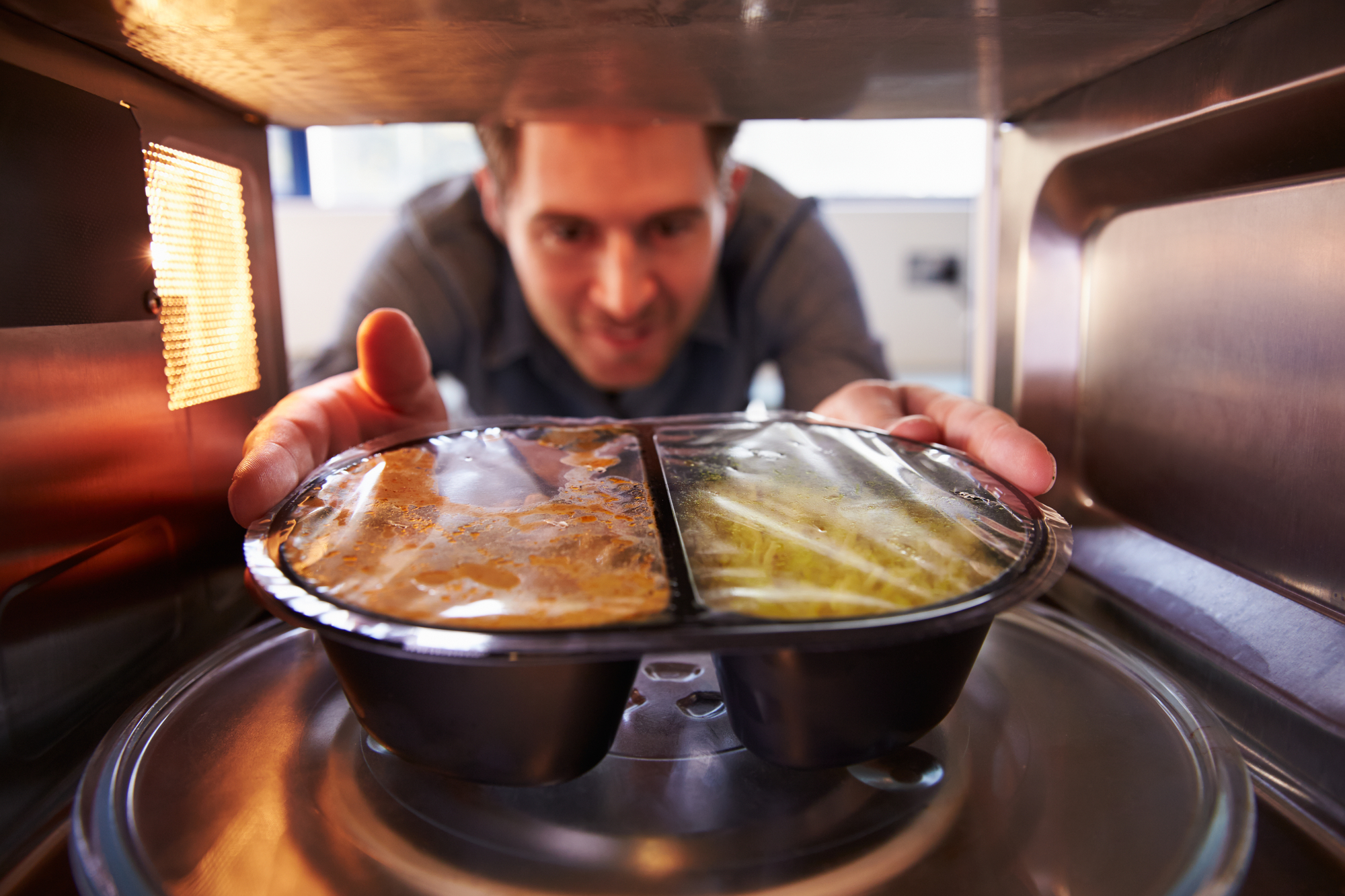 A person places a black plastic tray containing pre-packaged microwave meals into a microwave oven. The person is leaning forward and smiling, looking at the food through the open microwave door. The microwave is lit up and the food is ready for heating.