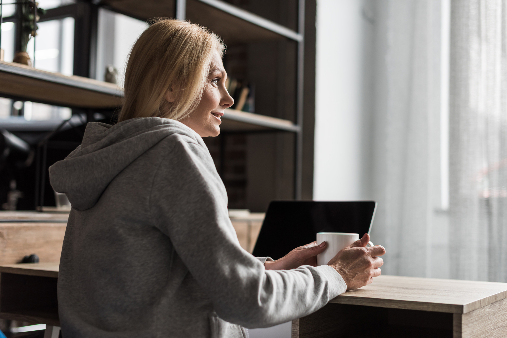 A woman with long blonde hair, wearing a grey hoodie, sits at a wooden desk holding a white mug. She looks out of a window with light streaming in. A laptop is on the desk in the background, with shelving and various items behind her.