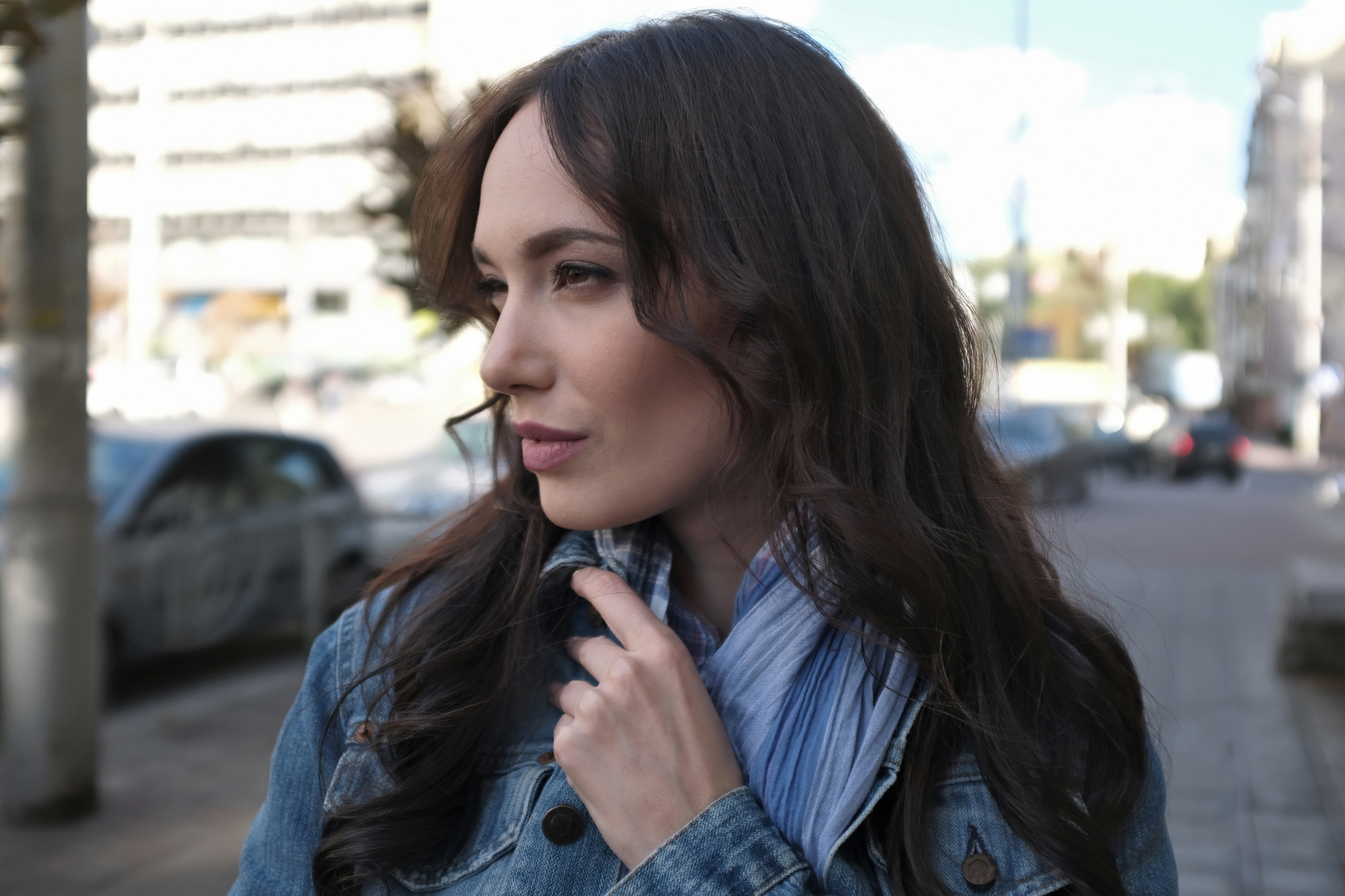 A person with long, dark hair stands outdoors in an urban setting. They are wearing a denim jacket and a light blue scarf, looking to the left with a thoughtful expression. The background is slightly blurred, showing buildings, trees, and a car.