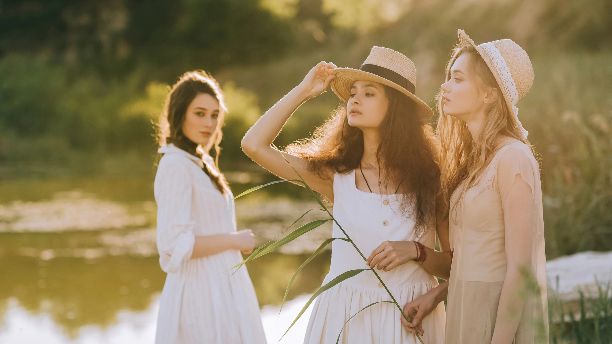 Three women in light, summery dresses and wearing straw hats stand in a sunlit, grassy outdoor setting near a body of water. One woman looks into the distance while holding her hat, another gazes ahead, and the third glances back over her shoulder.