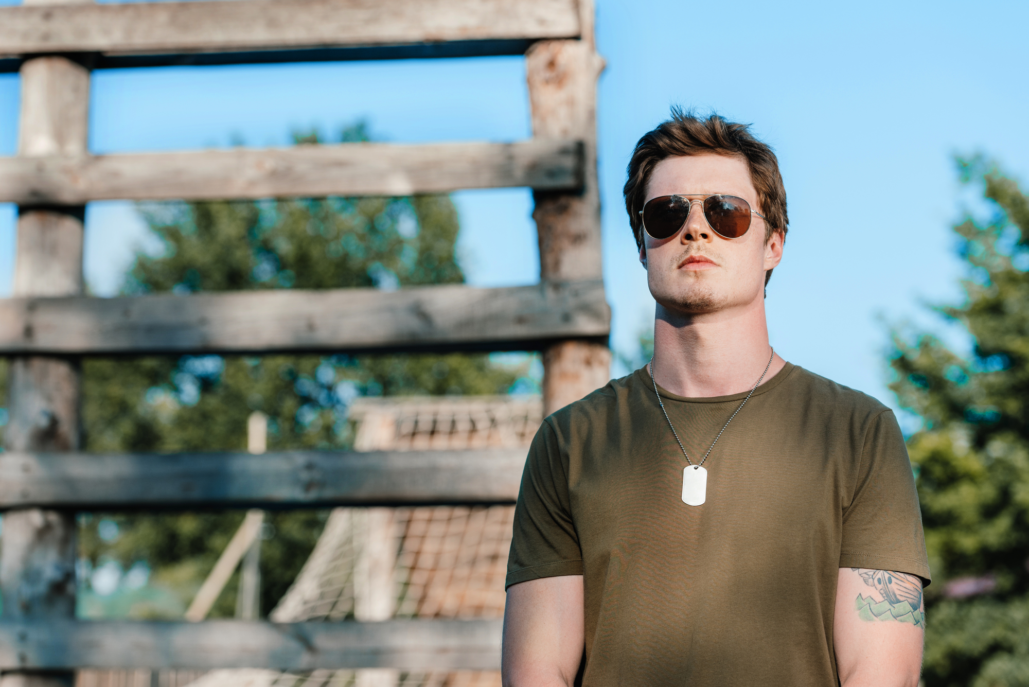 A man wearing sunglasses and a green t-shirt stands outdoors with a serious expression. He has a dog tag necklace and a tattoo on his left arm. The background features a rustic wooden structure and greenery under a clear blue sky.
