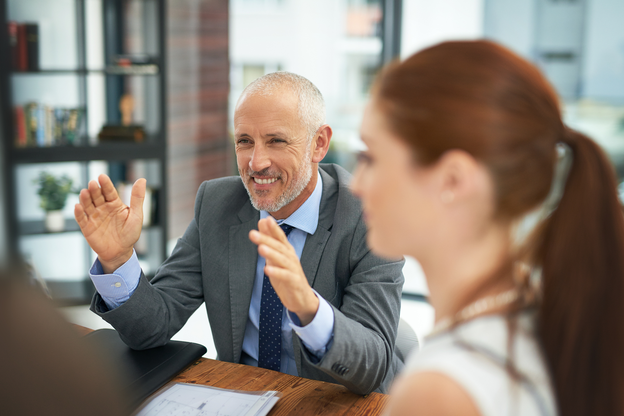 A man with gray hair and wearing a suit is smiling and gesturing with his hands while sitting at a wooden table in an office setting. A woman with red hair and wearing a white top is in the foreground, slightly out of focus. Bookshelves are visible in the background.