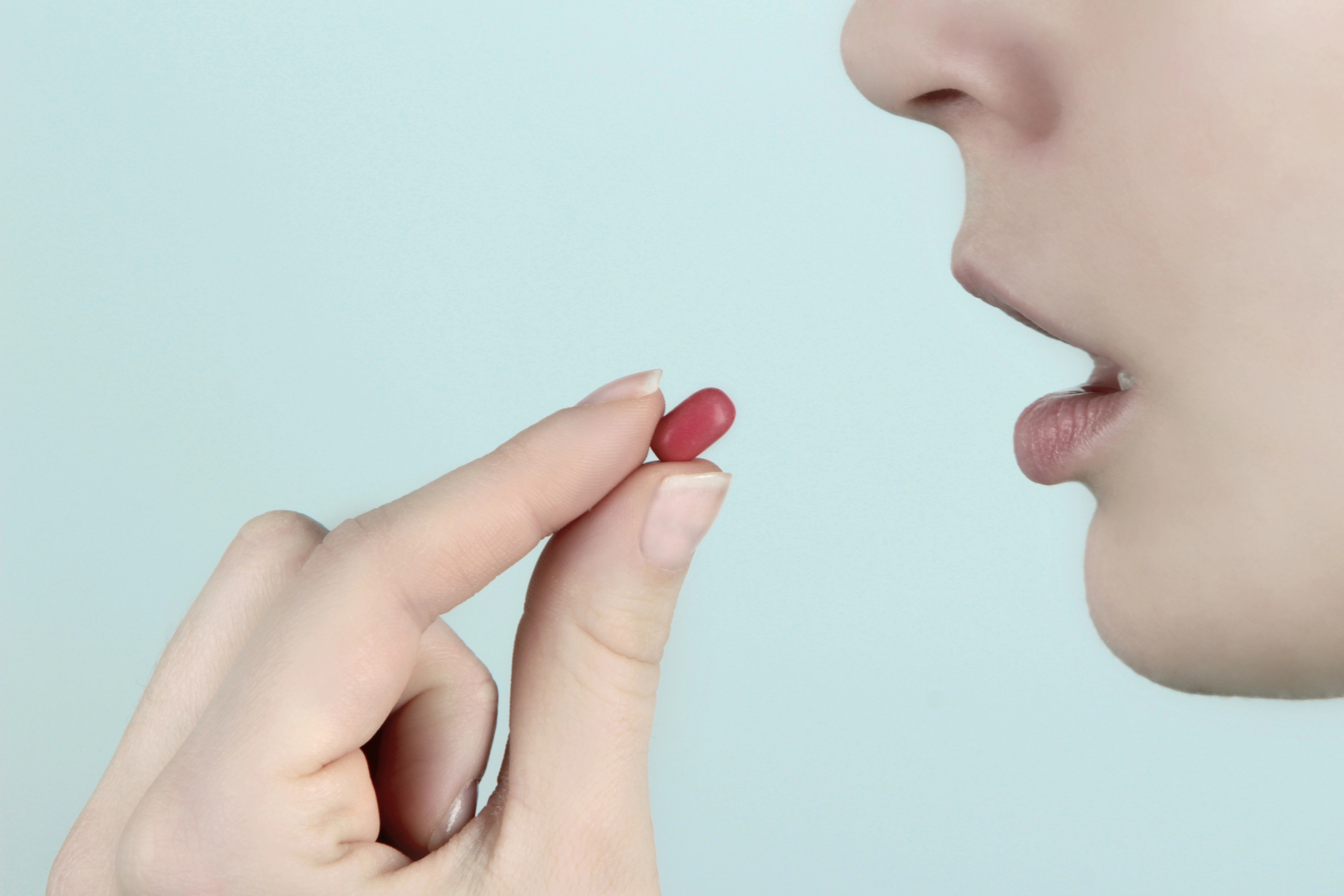 Close-up of a person about to take a red pill, which is held between the thumb and index finger of their right hand. The person's mouth is slightly open, and the background is a plain, light blue.