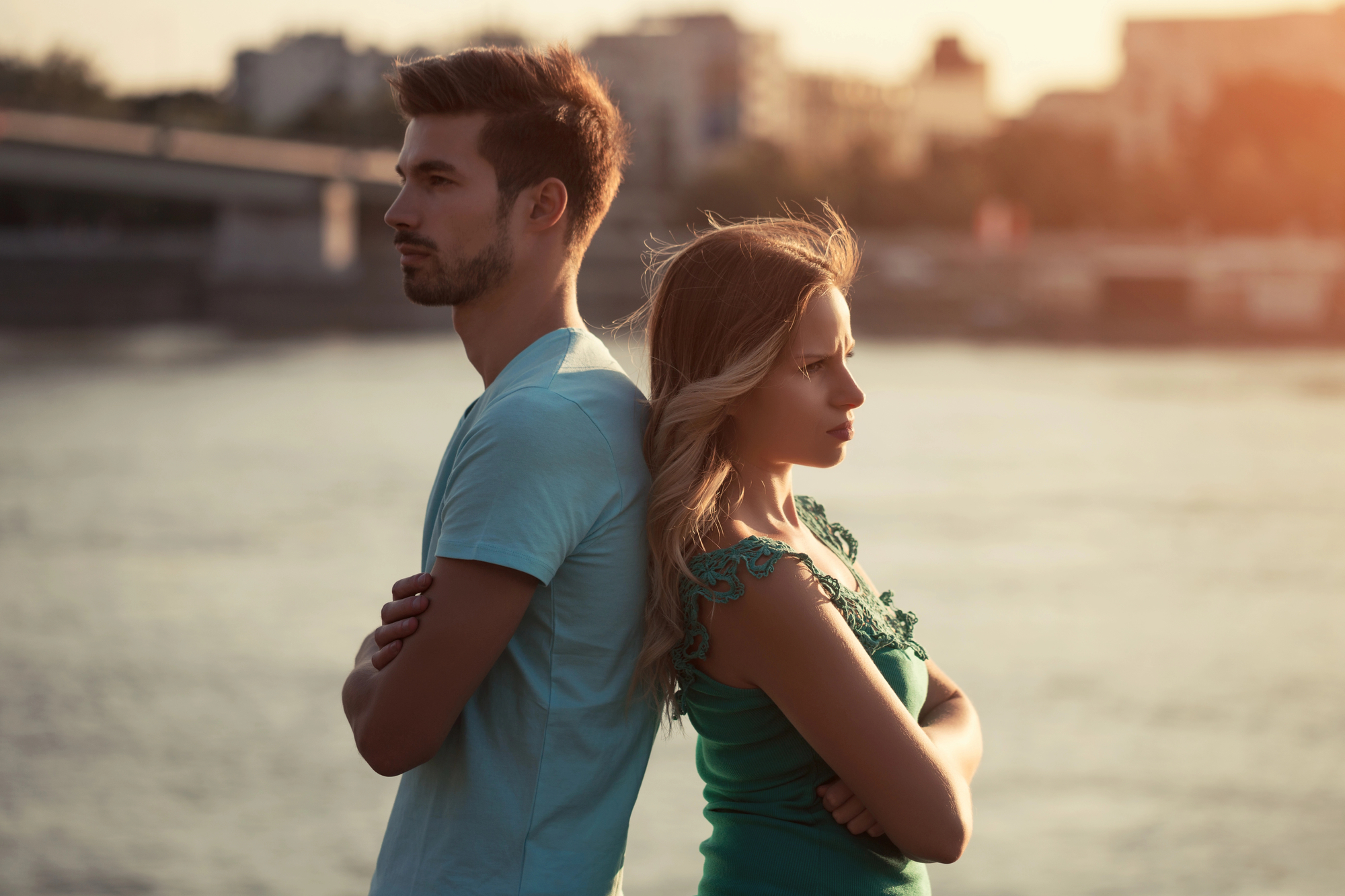 A man and woman stand back to back with arms crossed, looking away from each other. The background shows a waterfront with a bridge and buildings under a warm, late-afternoon light. Both appear to be deep in thought or upset.