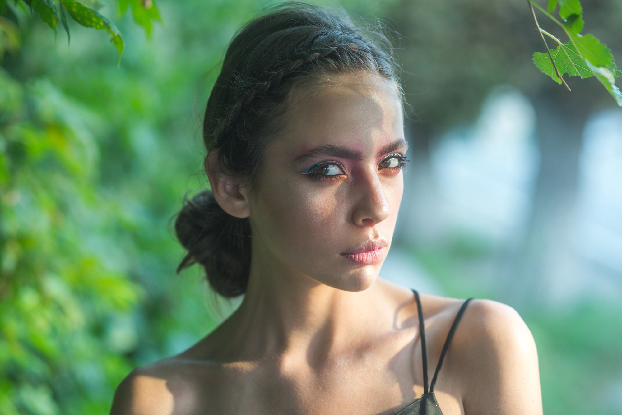A woman with braided dark hair looks intently at the camera. She wears subtle makeup with a touch of blush and pink eyeshadow. She has a sleeveless top and is surrounded by green foliage, with sunlight casting gentle shadows on her face.