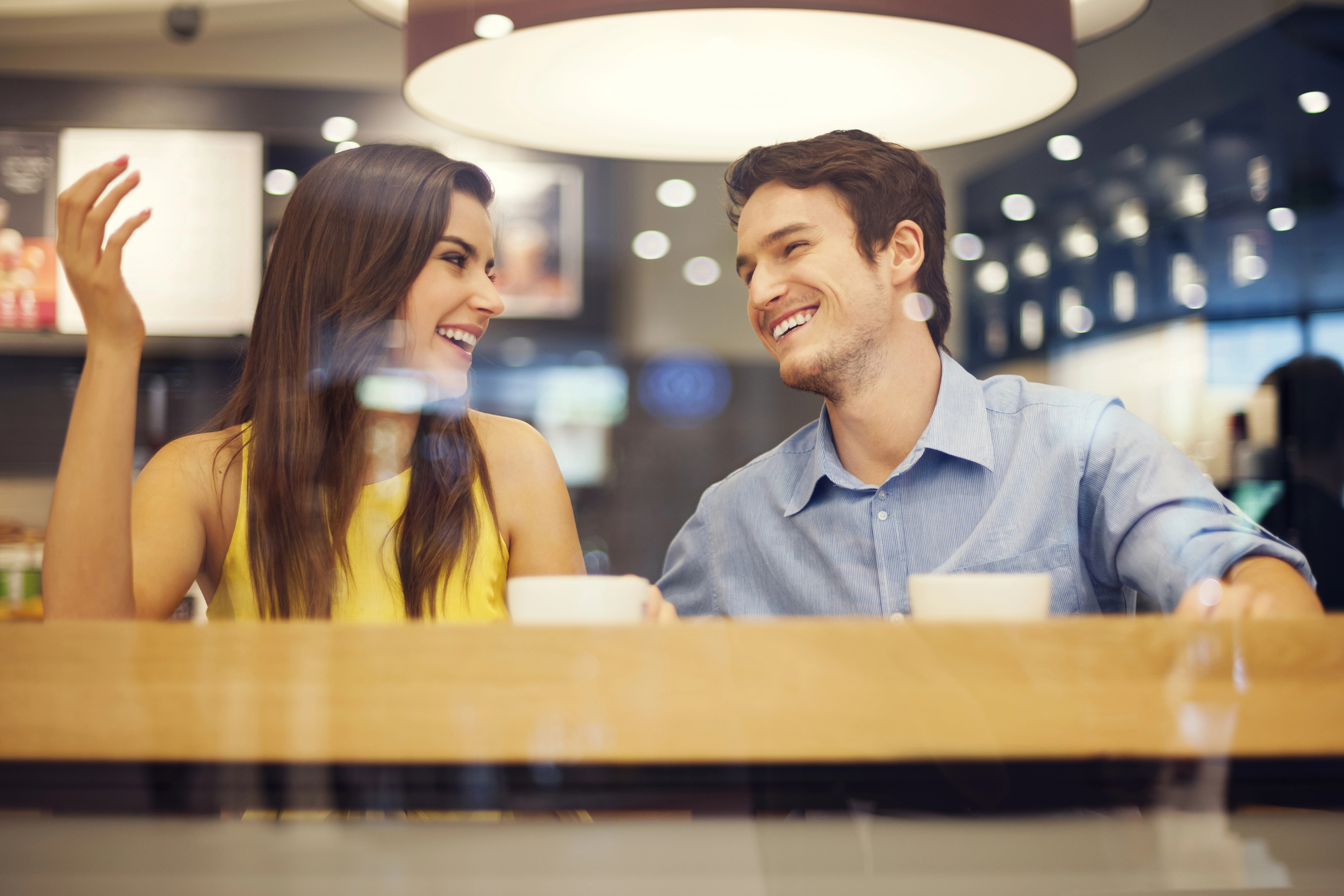 A woman and a man sit at a table in a café, smiling and engaging in conversation. The woman wears a yellow top, and the man wears a blue shirt. They appear to be enjoying each other's company against a backdrop of softly blurred café lights.