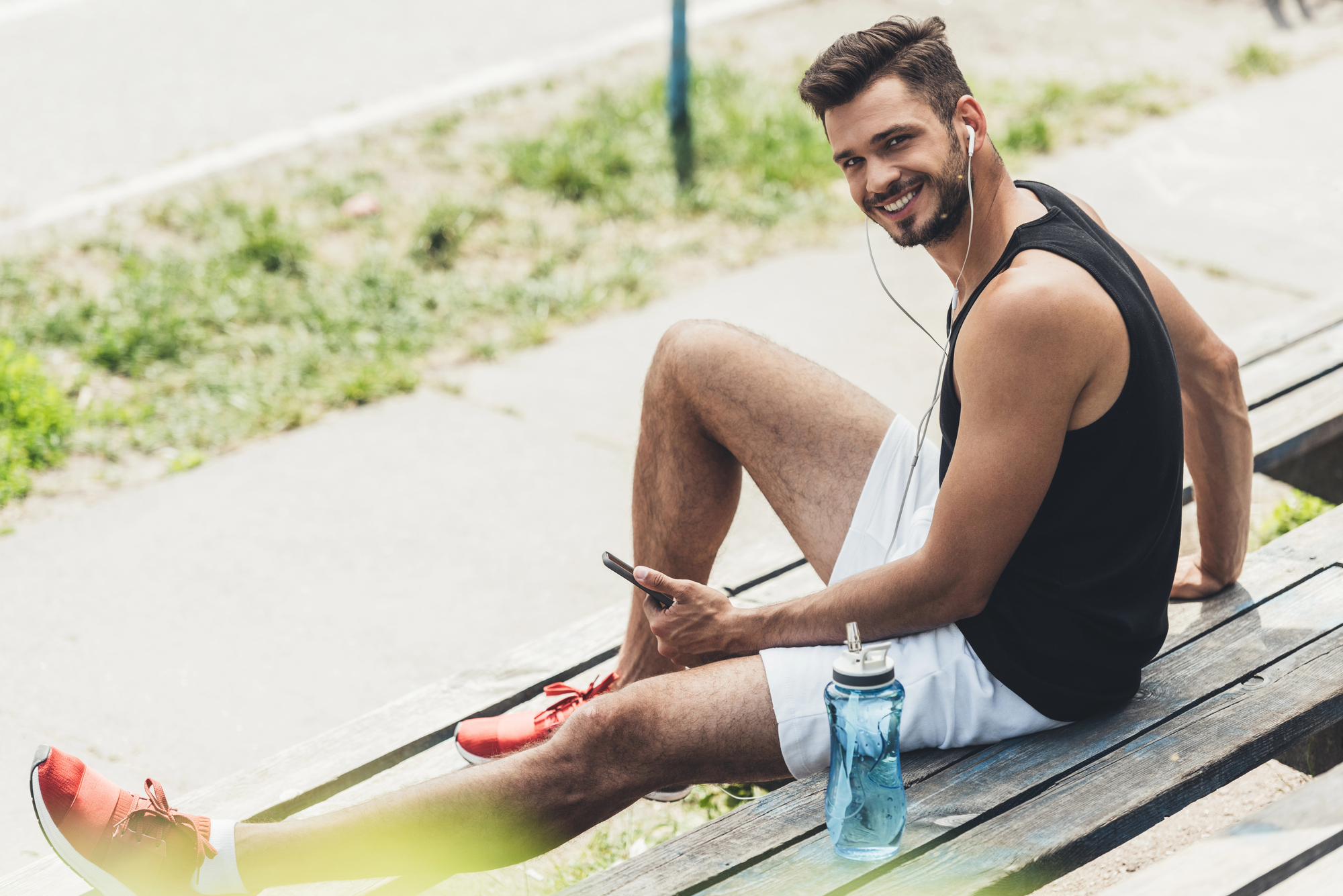 A young man sits on wooden bleachers, smiling while holding a smartphone in one hand and wearing earphones. He is dressed in a black tank top, white shorts, and red sneakers. A blue water bottle is placed on the bench next to him. The background shows a pathway and greenery.