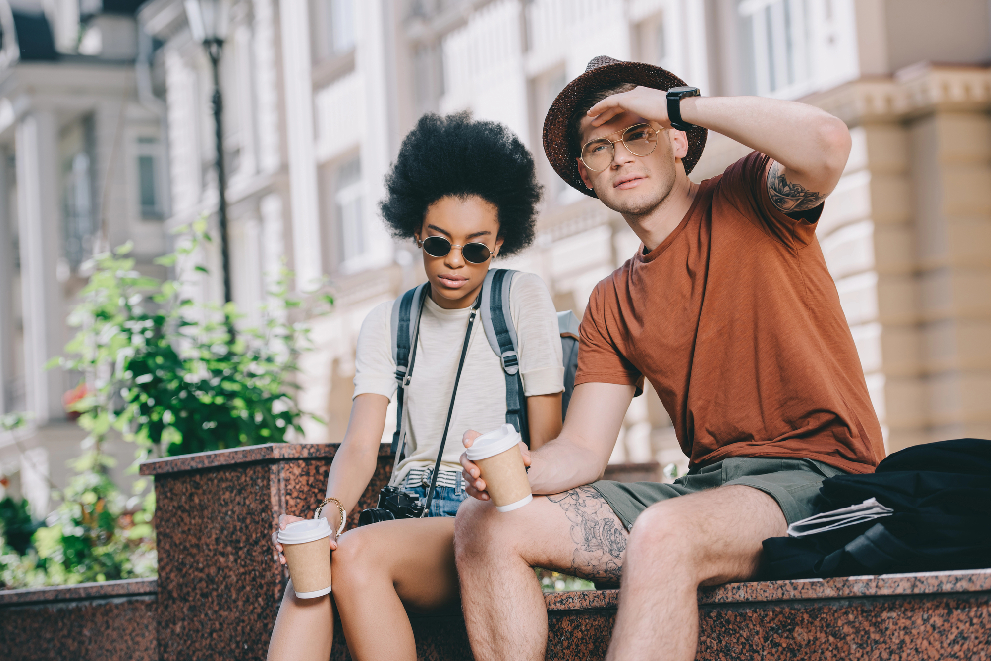 Two friends sit on a ledge outdoors, each holding a coffee cup. The person on the left wears sunglasses, a backpack, and casual clothes, looking down at their coffee. The person on the right, also in casual attire and wearing a hat and sunglasses, shields their eyes from the sun.