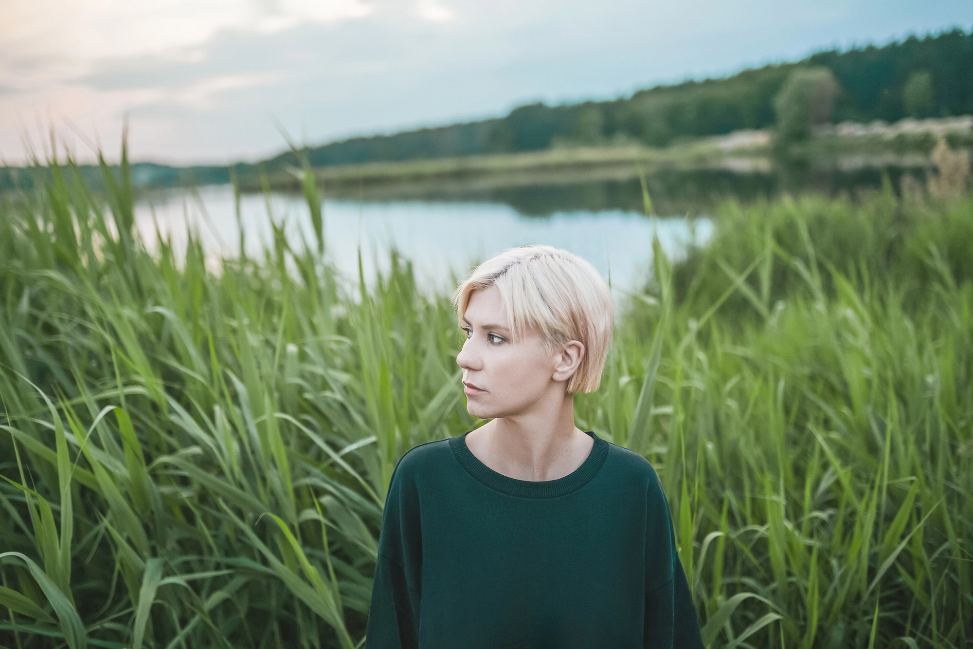 A person with short, light blonde hair stands amidst tall green grass by a serene lake. They are wearing a dark green top and looking to the side. The background consists of the calm water of the lake and a treeline under a partly cloudy sky.