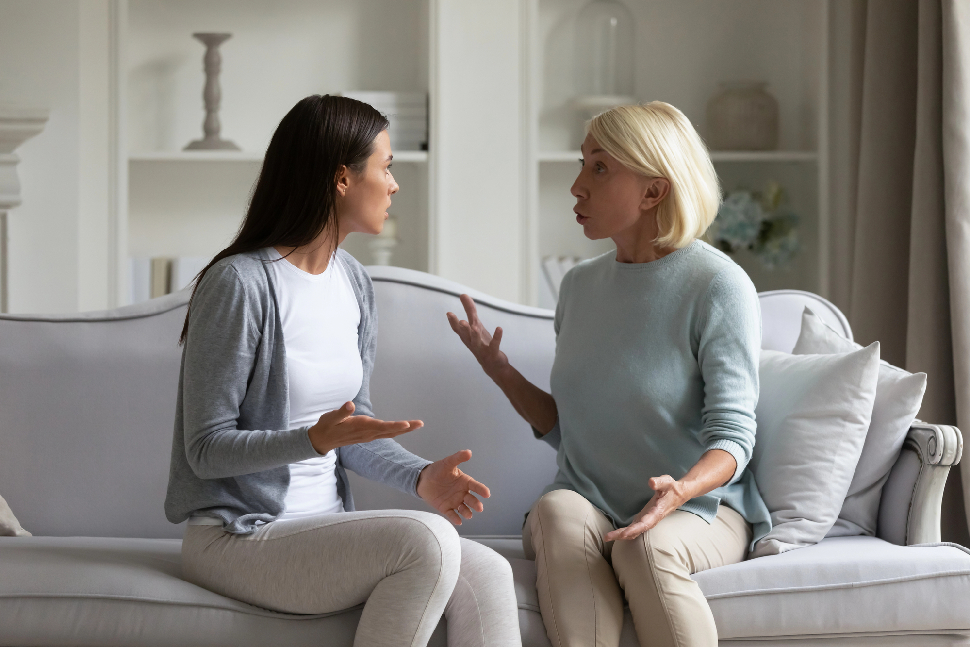 Two women, one younger with dark hair and one older with blonde hair, are sitting on a beige couch in a living room. They are engaged in a heated discussion, with both gesturing emphatically. Light-filled shelves and decorations are visible in the background.