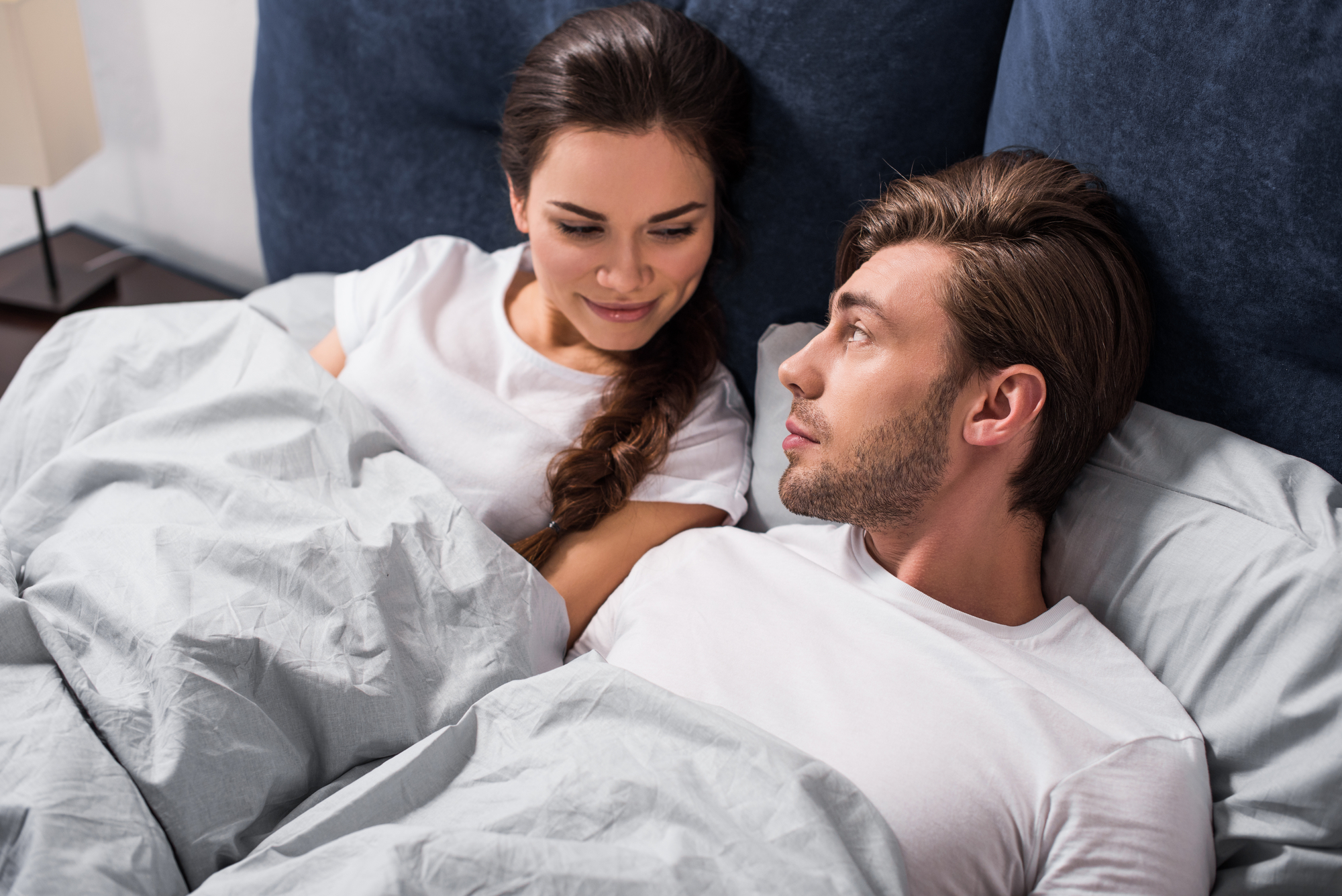 A man and woman lie in bed together under grey blankets, both wearing white shirts. They face each other, the woman smiling softly while the man gazes at her. The background is dark blue with a nightstand visible. The atmosphere appears calm and intimate.