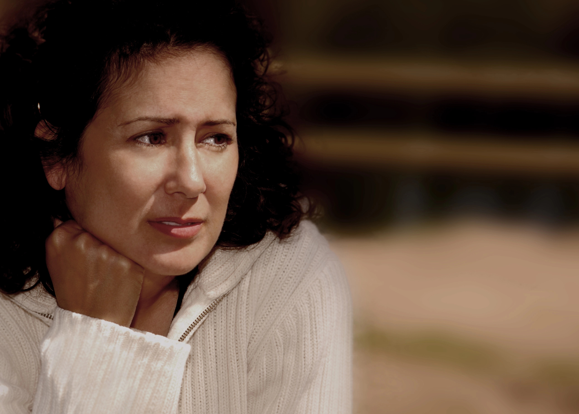 A woman with curly dark hair is leaning her head on her hand, looking contemplative. She is wearing a white sweater and has a thoughtful expression on her face. The background is softly blurred, putting the focus on her face and emotions.