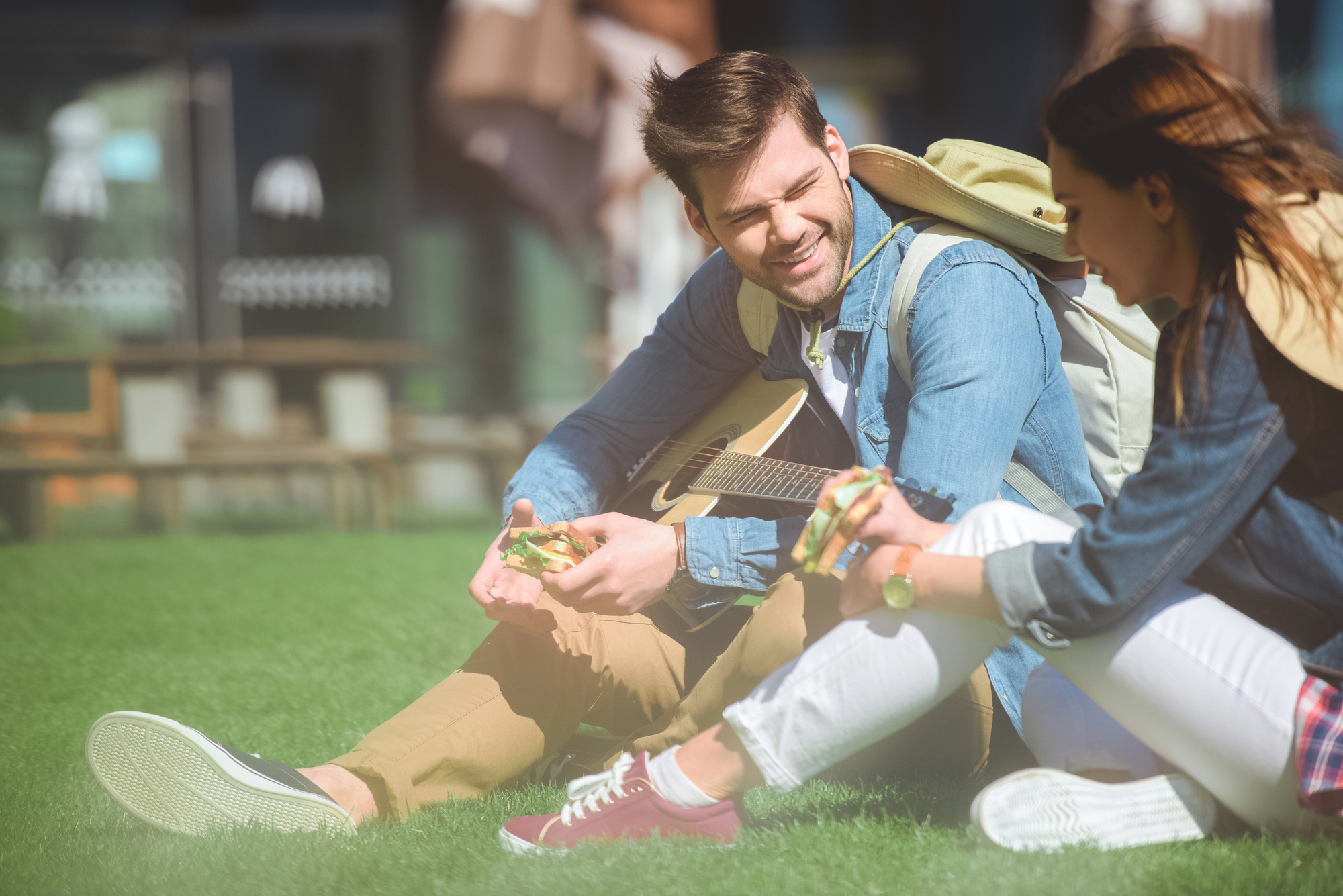 A man and woman sit on the grass, enjoying snacks. The man holds a guitar and dons a backpack, while both wear casual clothing. They are outdoors, possibly in a park, and in a relaxed, cheerful conversation.