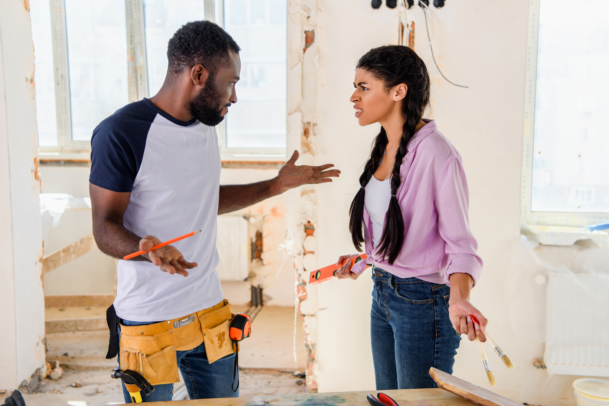 A man and woman are arguing in a partially renovated room. The man has a pencil in hand and is wearing a tool belt, while the woman, holding a paintbrush and measuring tape, looks frustrated. Both are standing near a table with renovation tools.