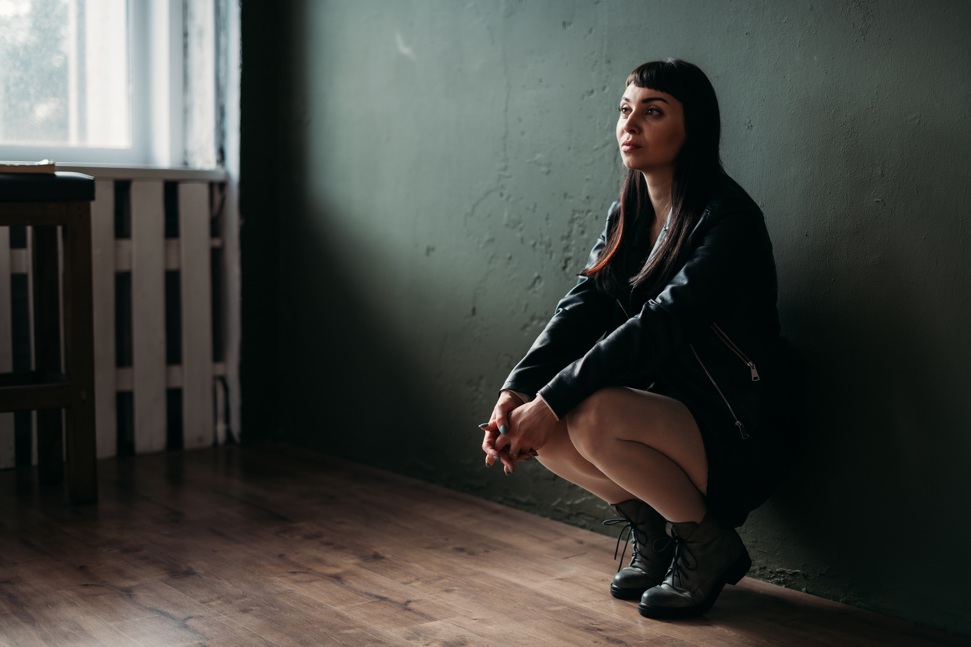 A woman with long dark hair sits on her heels against a dark green wall in a dimly lit room with wooden floors. She is wearing a black dress and jacket, with her hands resting on her knees, looking contemplative. A window with diffused light is in the background.