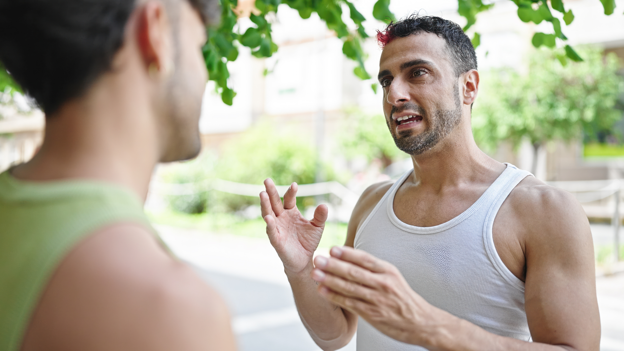 Two men are engaged in a conversation outdoors under a tree with green leaves. One man, facing the camera, gestures with his hands while speaking. Both are wearing sleeveless tops and appear to be in a lively discussion. The background is blurred with greenery.