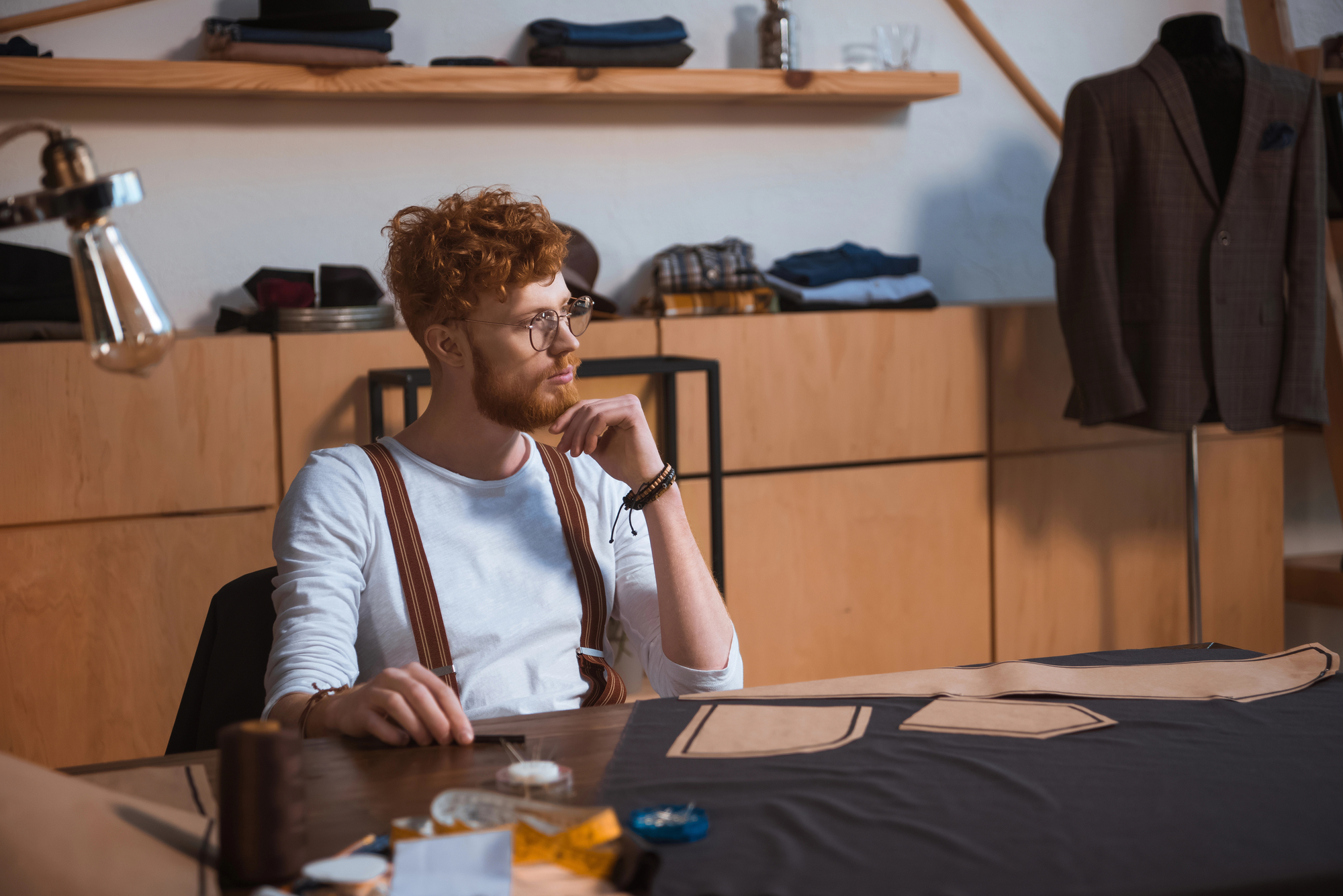 A person with curly red hair and glasses sits at a table in a tailor shop, surrounded by sewing materials and pattern pieces. They are wearing a white shirt and suspenders, gazing thoughtfully to the side. Clothing and fabric are visible in the background.