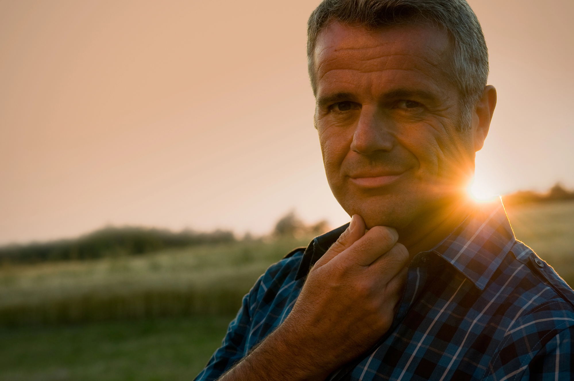 A man with short gray hair and a slight smile stands outdoors in a field at sunset. He is wearing a blue and black checkered shirt and touches his chin with his left hand. The sun is behind him, casting a warm glow. Trees and a grassy field are visible in the background.
