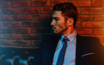A man in a dark suit and blue tie sits on a black leather sofa. He has neatly styled hair and a trimmed beard. The background is a brick wall with red and blue lighting creating a dramatic contrast. The man looks pensively to his left.