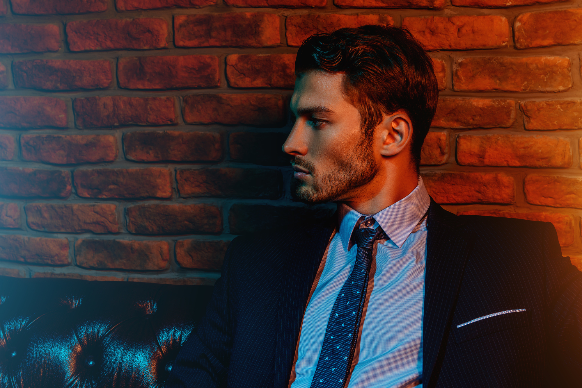 A man in a dark suit and blue tie sits on a black leather sofa. He has neatly styled hair and a trimmed beard. The background is a brick wall with red and blue lighting creating a dramatic contrast. The man looks pensively to his left.