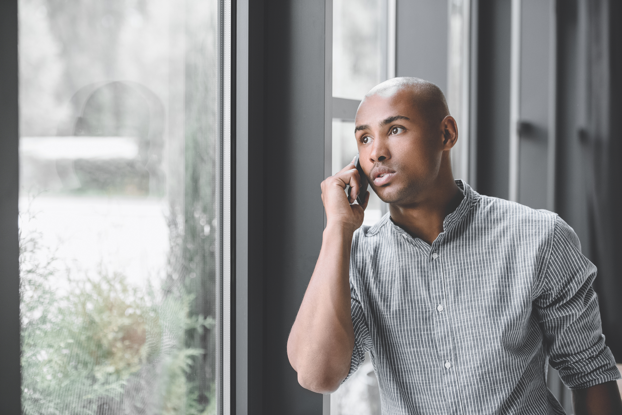A man in a grey checkered shirt is standing by a large window, looking outside while talking on a smartphone. His expression is focused and contemplative, suggesting he is engaged in an important conversation. The background is lightly blurred.