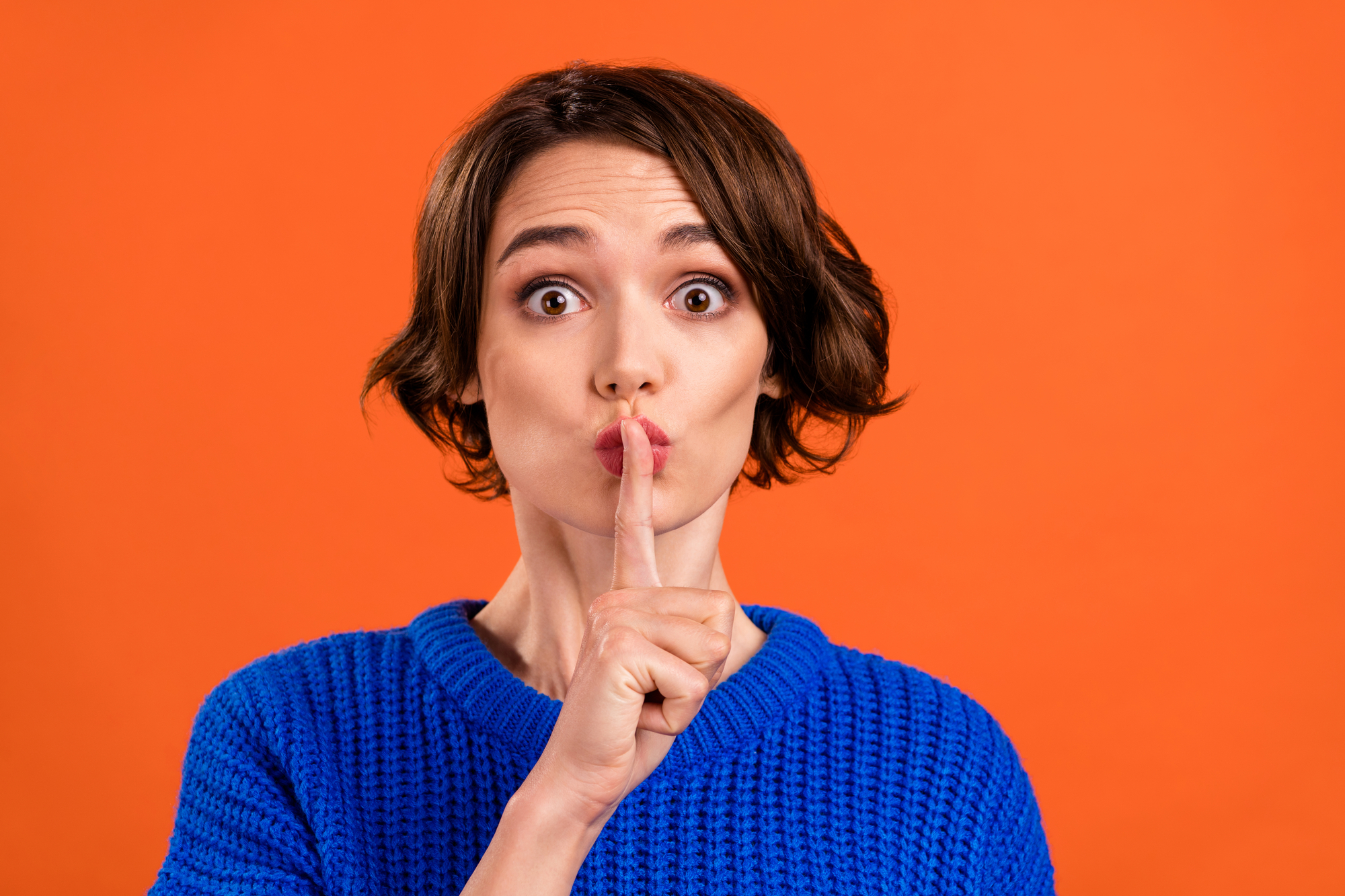 A person with short brown hair is standing against an orange background, wearing a blue sweater. They are looking directly at the camera with wide eyes, holding their index finger up to their lips in a gesture signaling to be quiet.