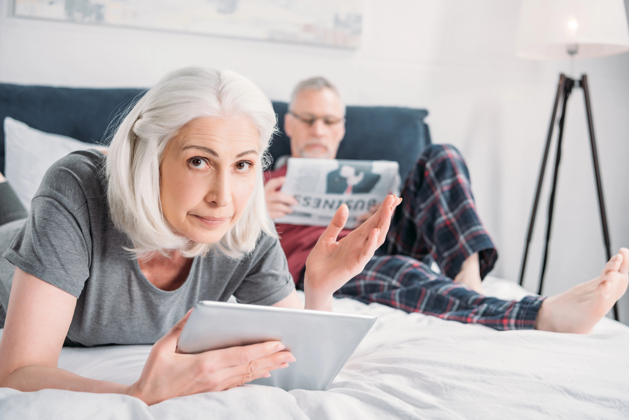 An older woman with gray hair lies on a bed holding a tablet, looking at the camera with a raised eyebrow and hand gesture. In the background, an older man with gray hair and glasses, wearing plaid pajama pants, sits on the bed reading a newspaper.