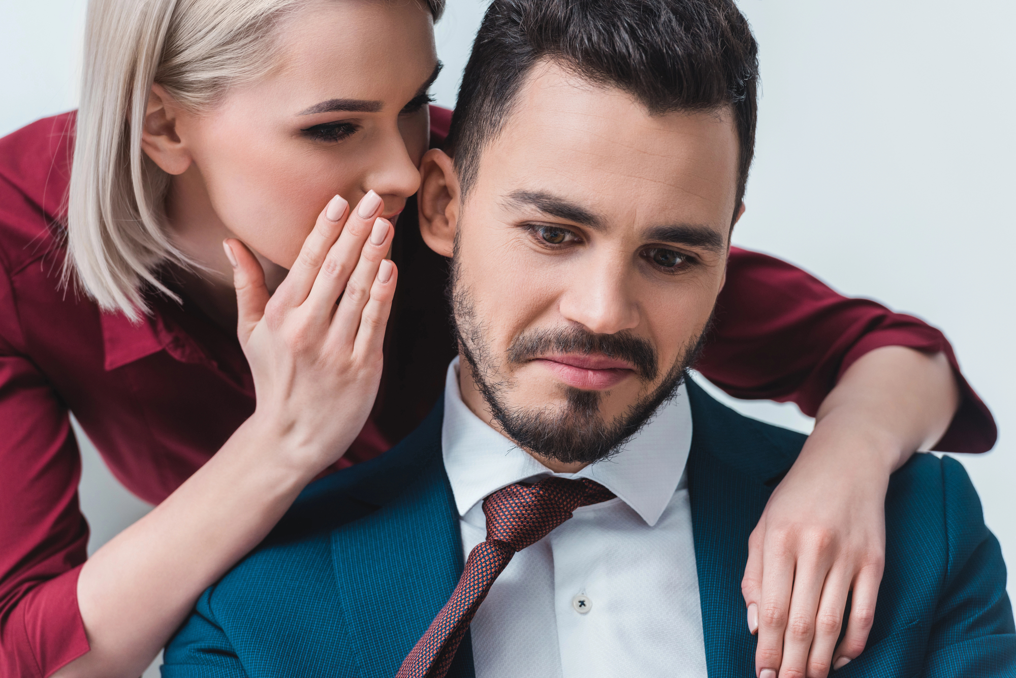 A woman with blonde hair, wearing a red shirt, leans in to whisper into the ear of a man with dark hair and a beard, who is wearing a blue suit and red tie. The man has a slight frown and is looking downwards. Both appear to be against a plain white background.