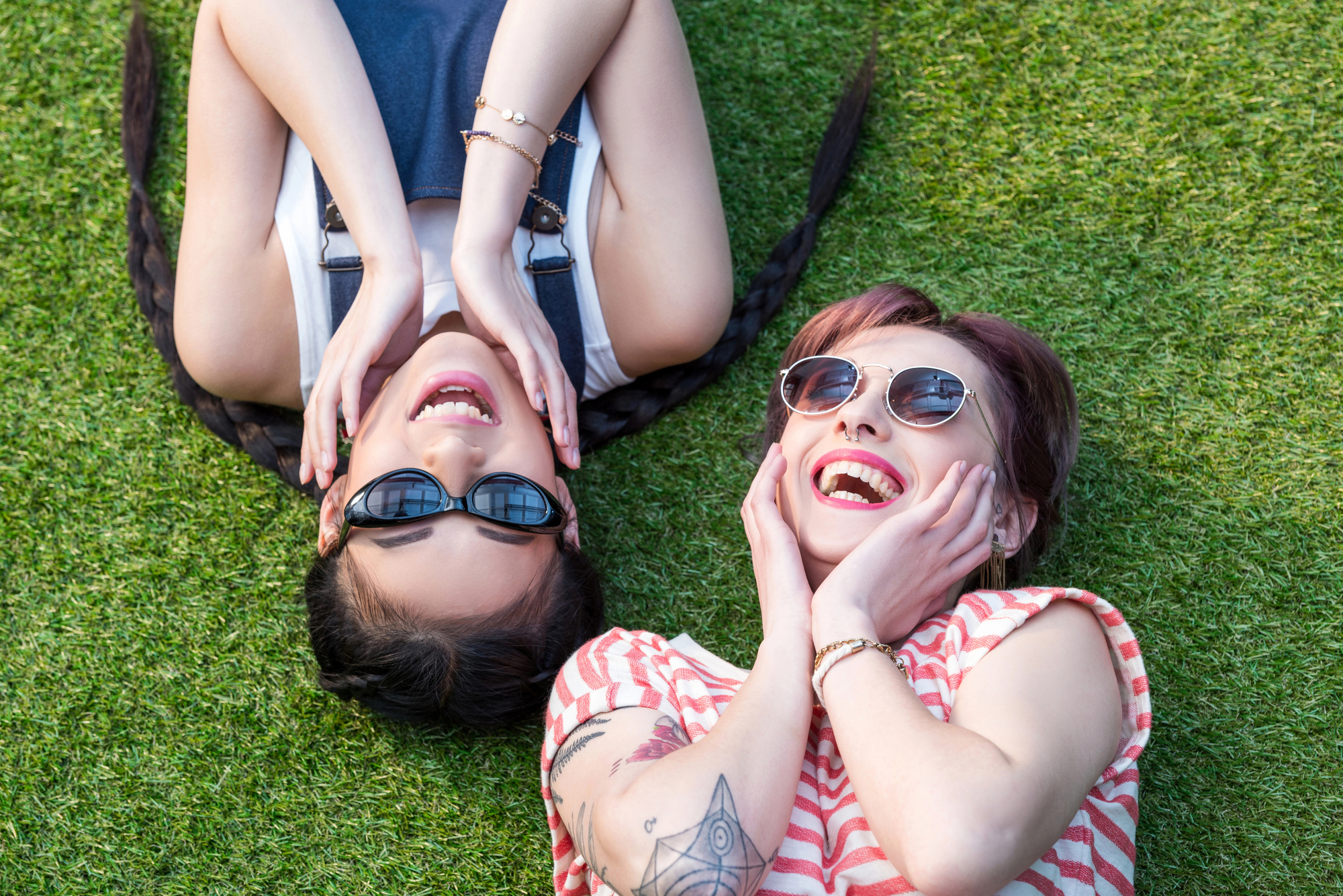Two friends lie on the grass, heads touching and hands on their cheeks. Both wear sunglasses and smile brightly. One wears a striped shirt and has tattoos while the other has a sleeveless top and braided hair. The mood is joyful and carefree.