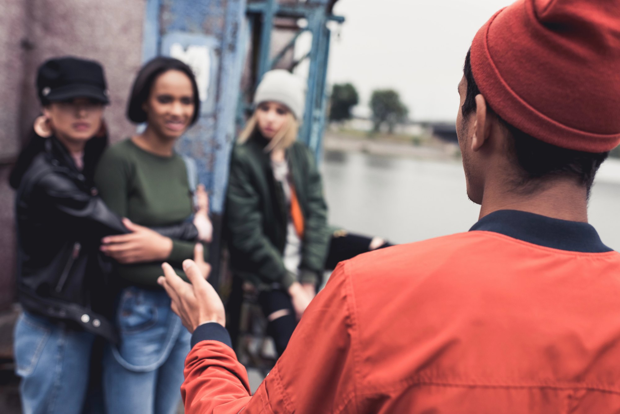 Four young adults are outdoors near a body of water. One person in the foreground is wearing a red jacket and beanie, gesturing as they talk to the other three, who are standing and sitting, listening intently. Their clothing suggests a casual, cool weather setting.