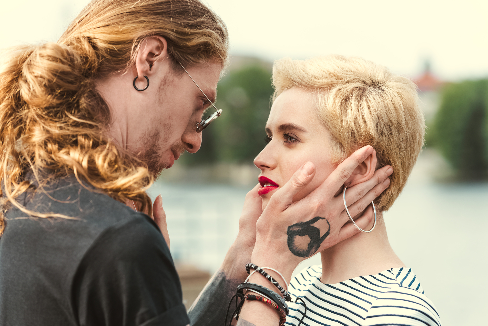 A couple gazes into each other's eyes with intense emotion. The man with long hair holds the woman's face tenderly. The woman has short blonde hair and bright red lipstick. They are standing outdoors, with blurred greenery and water in the background.