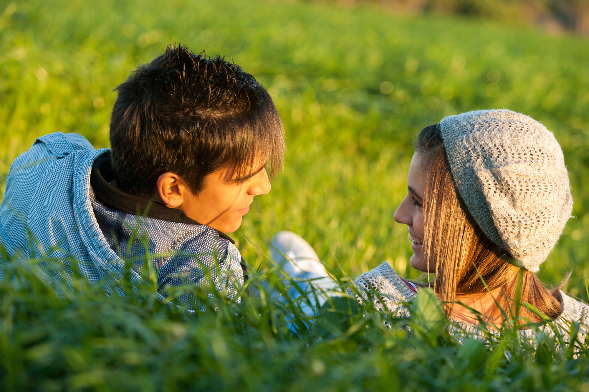 A young couple lies in a grassy field, facing each other and smiling warmly. The person on the left has short dark hair, while the person on the right wears a grey beanie and has long hair. The sunlight casts a soft, golden glow over them and the lush green grass.
