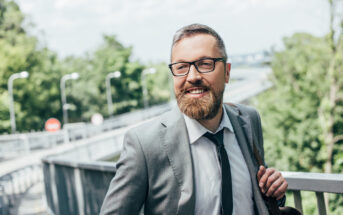 A bearded man with glasses wearing a gray suit and black tie stands smiling on a bridge in an outdoor setting with green foliage and a bright sky in the background.