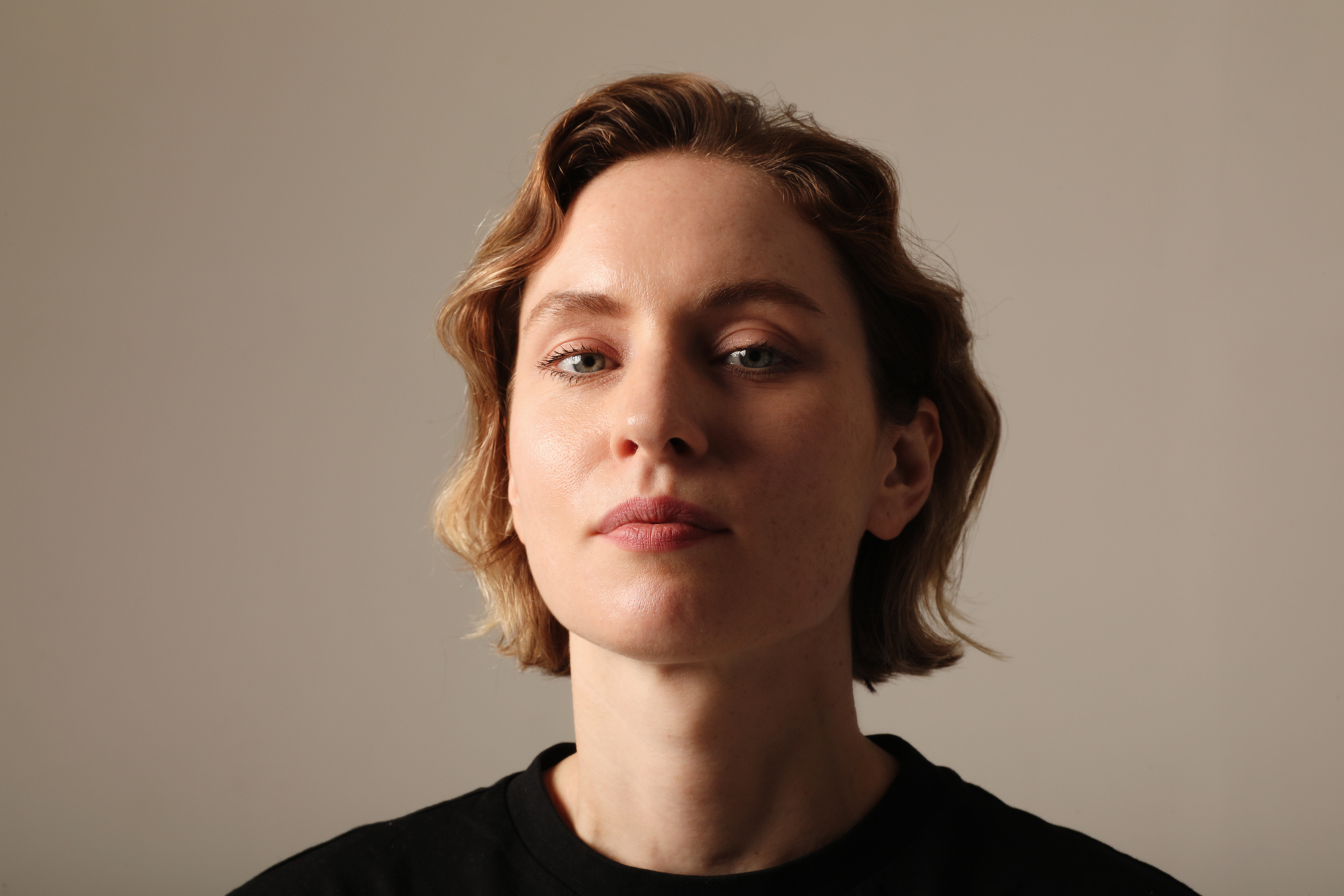 A person with short, wavy, light brown hair is looking directly at the camera with a neutral expression. They are wearing a black top and the background is plain and light-colored. Their skin appears to be fair, and their eyes seem calm and focused.