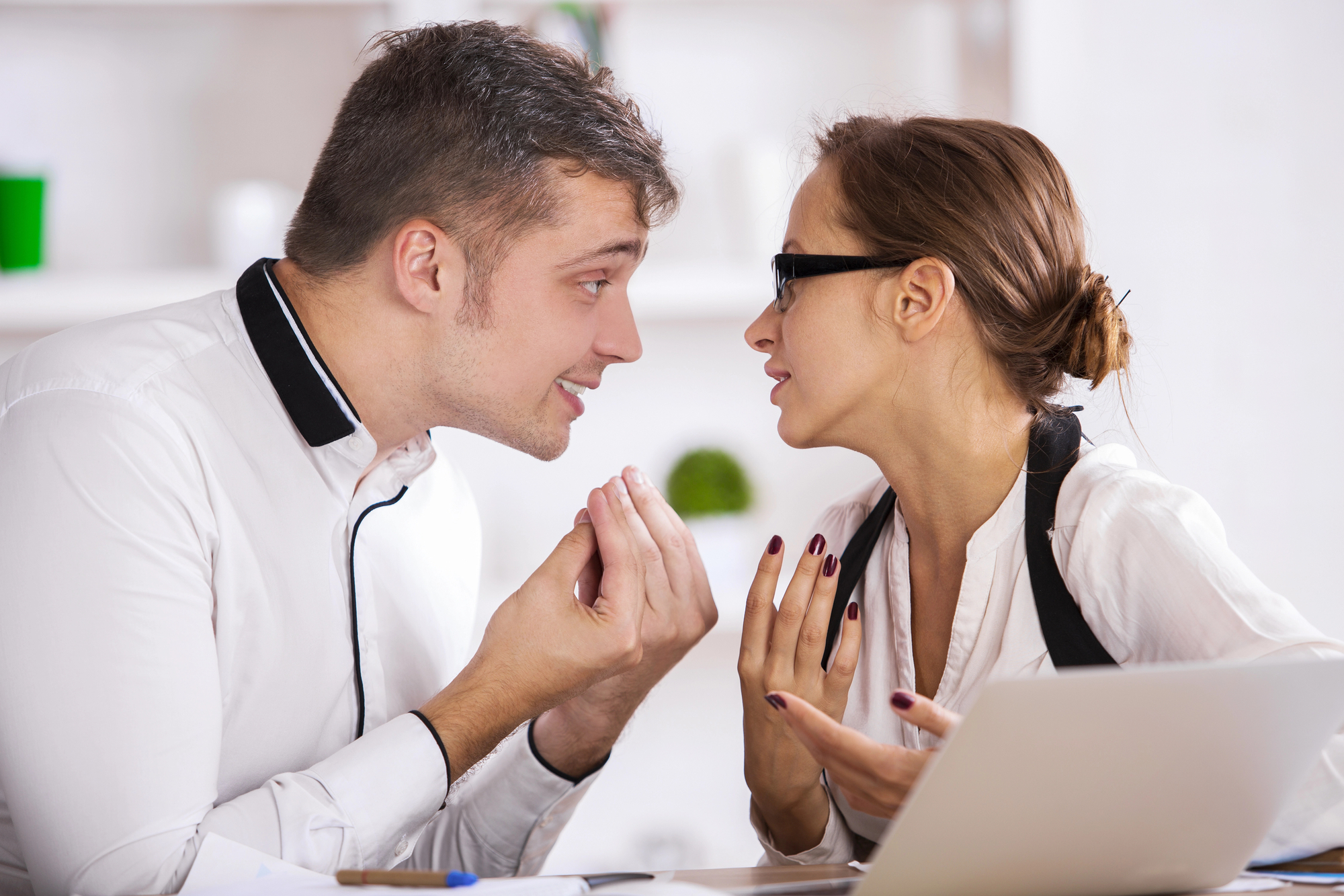 A man and a woman in a heated discussion at a desk in an office. The man is wearing a white shirt and gesturing with his hands, leaning towards the woman. The woman, also in a white shirt and wearing glasses, is responding with an intense expression. A laptop is open in front of them.