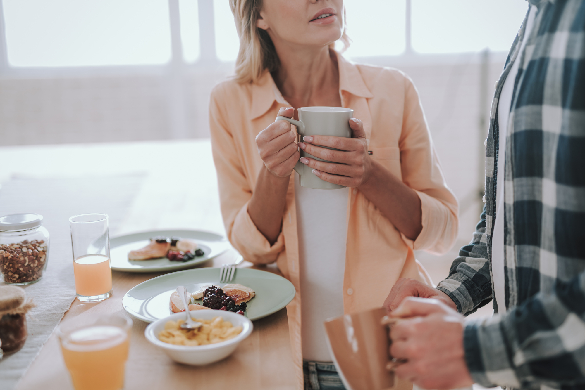 Two people are standing at a dining table, each holding a mug. On the table, there are plates with breakfast food, including pancakes with berries, a bowl of cereal, a glass of orange juice, and various jars. The lighting is bright and natural from nearby windows.