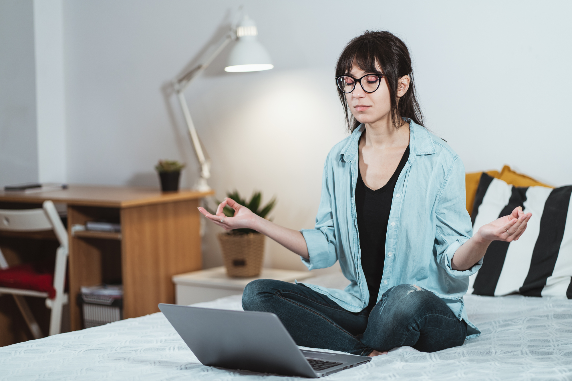 A person with glasses sits cross-legged on a bed, meditating with their eyes closed and hands in a mudra position. A laptop is open in front of them. The background includes a desk, a chair, a lamp, and a potted plant.