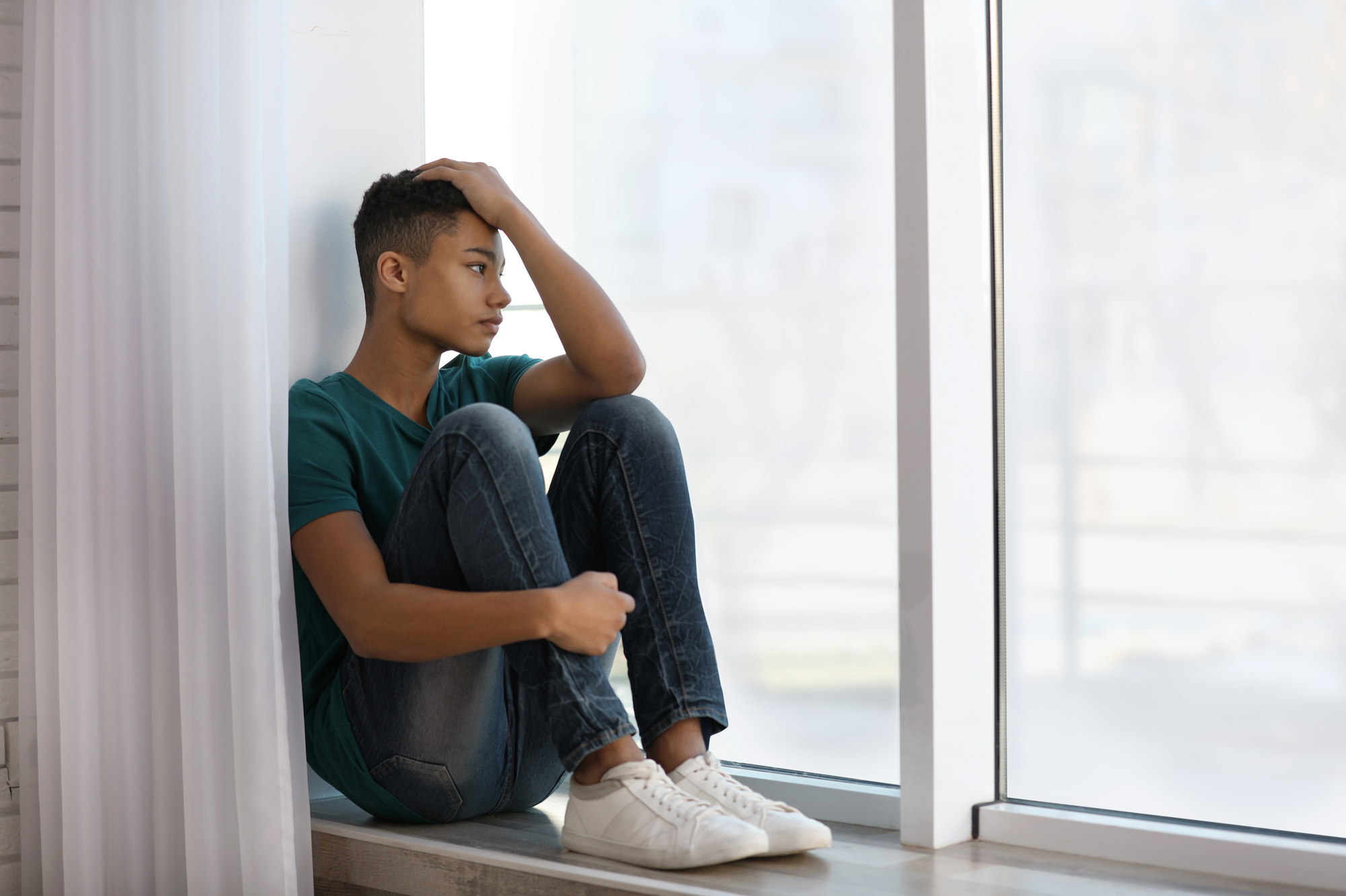 A young person sits on a windowsill, staring thoughtfully outside. They have short hair, wear a green t-shirt, jeans, and white sneakers. One arm rests on their knee, while the other hand touches their head. Light pours in from the large window beside them.