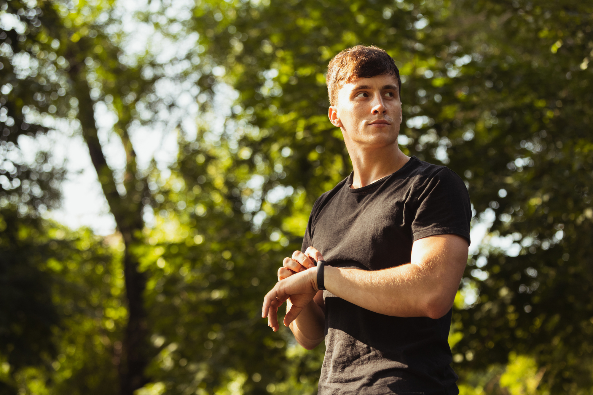 A young man with short brown hair stands outdoors, wearing a black T-shirt and a smartwatch. He is looking to his left with a thoughtful expression, set against a backdrop of lush, green trees and sunlight filtering through the leaves.