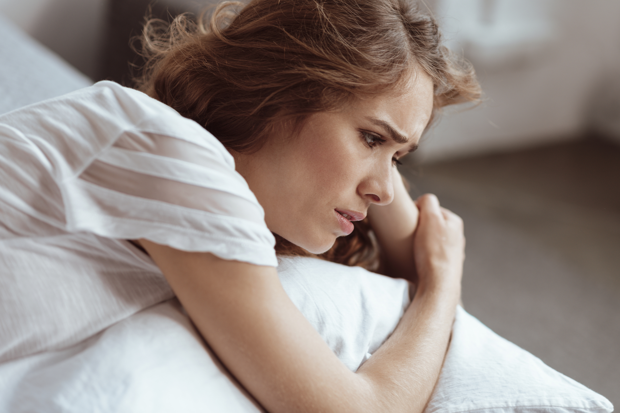 A woman with long, wavy hair and a white shirt leans on a pillow, looking pensive and worried. She gazes into the distance, her expression showing concern. Soft lighting fills the room, creating a calm yet somber atmosphere.