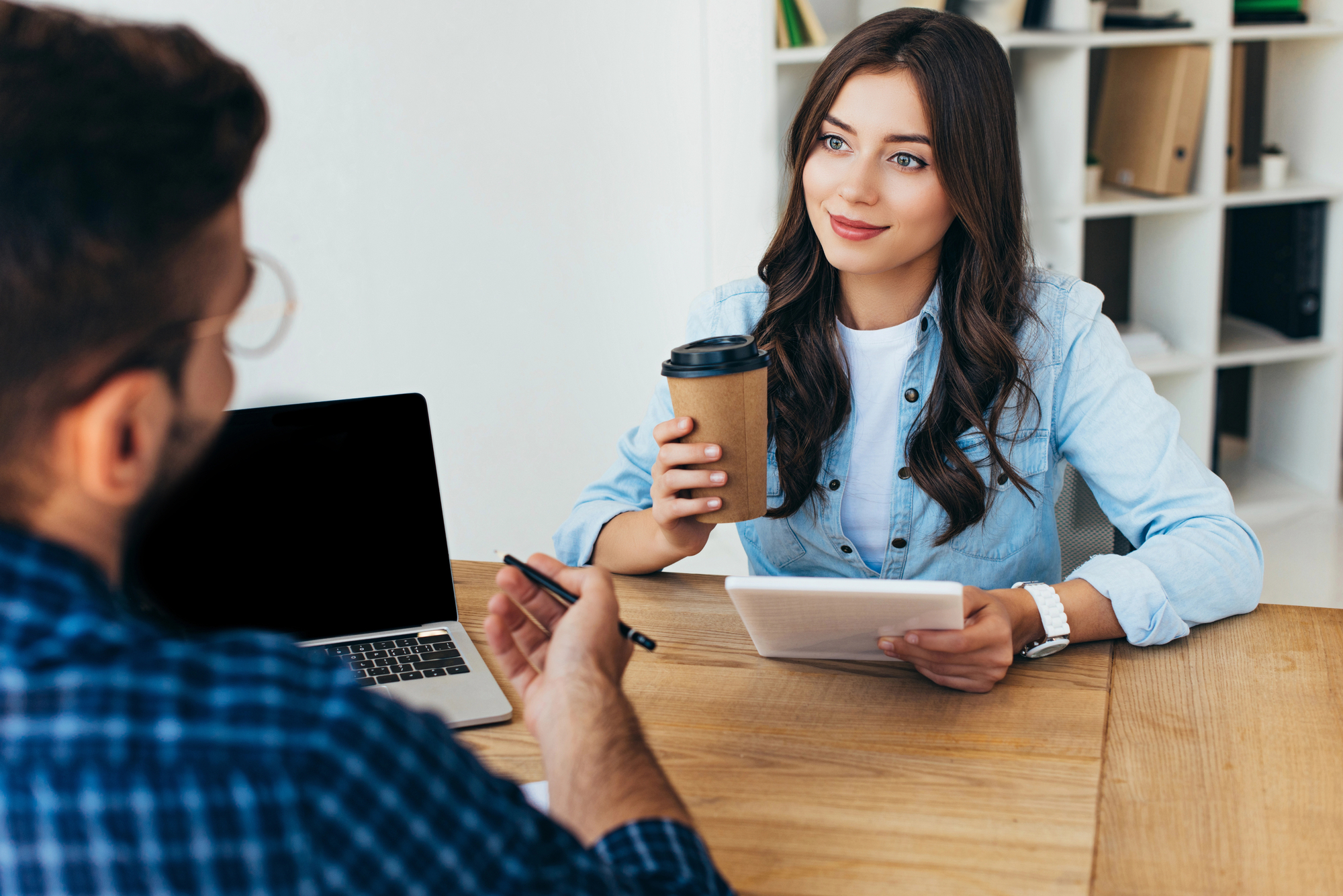 A woman with long brown hair, wearing a denim shirt, is sitting at a wooden table holding a takeaway coffee cup and a tablet, smiling at a man sitting opposite her. The man is in a plaid shirt, holding a pen. A laptop and bookshelf are in the background.