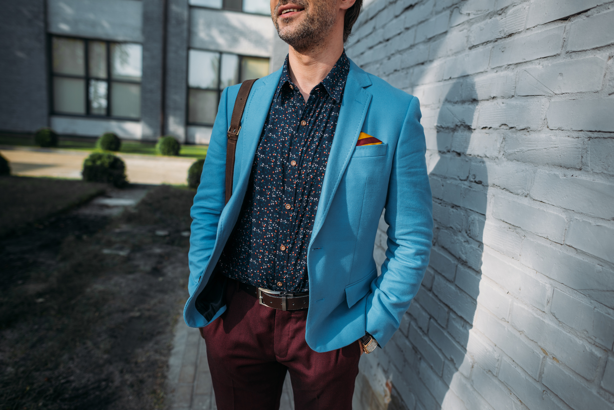A man stands confidently beside a white brick wall with one hand in his pocket. He's wearing a vibrant blue blazer, a dark patterned shirt, maroon pants, and a shoulder bag strap over his shoulder. A yellow pocket square peeks out from his blazer pocket.