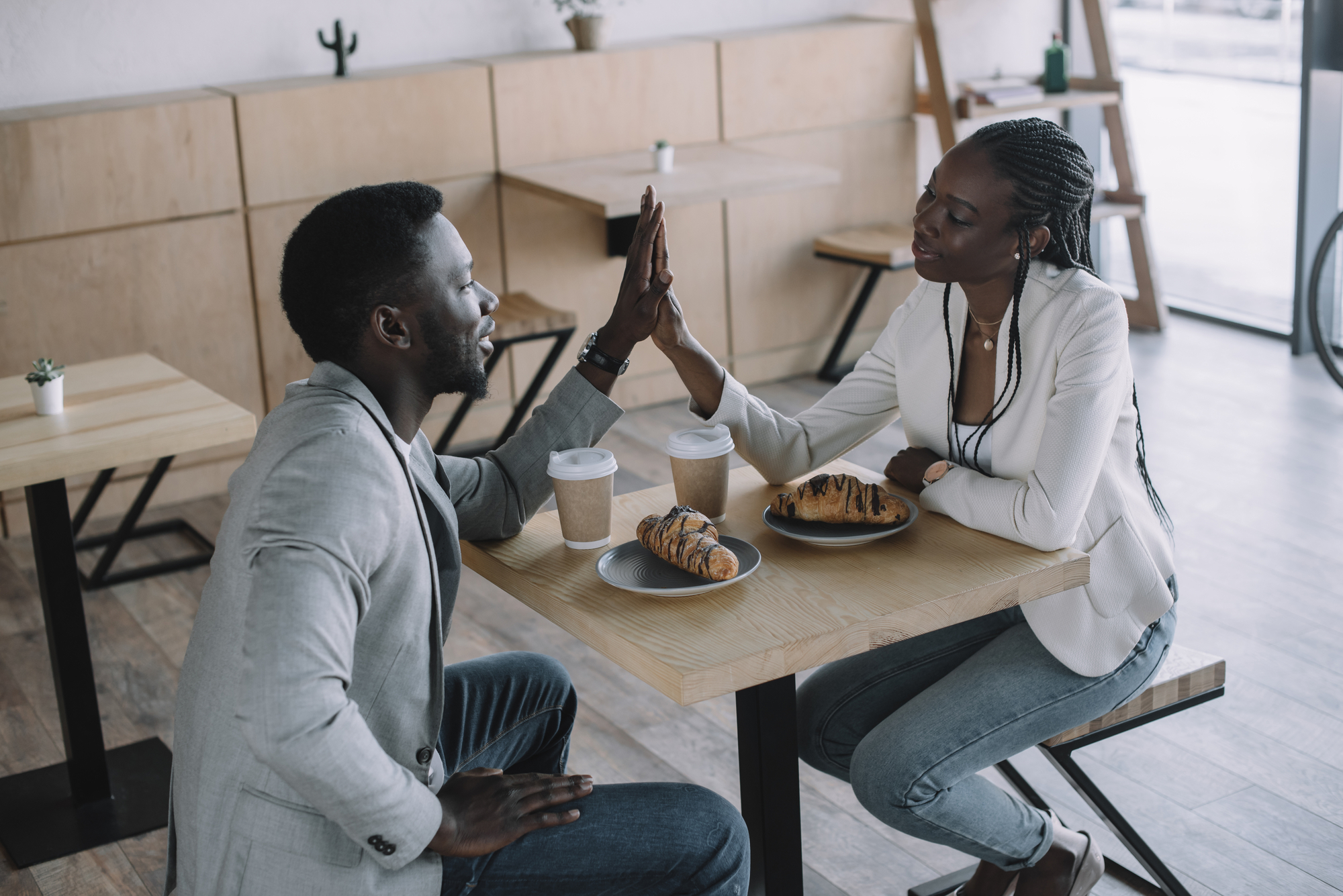Two people, a man and a woman, sit across from each other at a café table, smiling and giving each other a high-five. They have coffees and pastries on the table, and the café has a minimalist wooden interior with small plants on the tables.