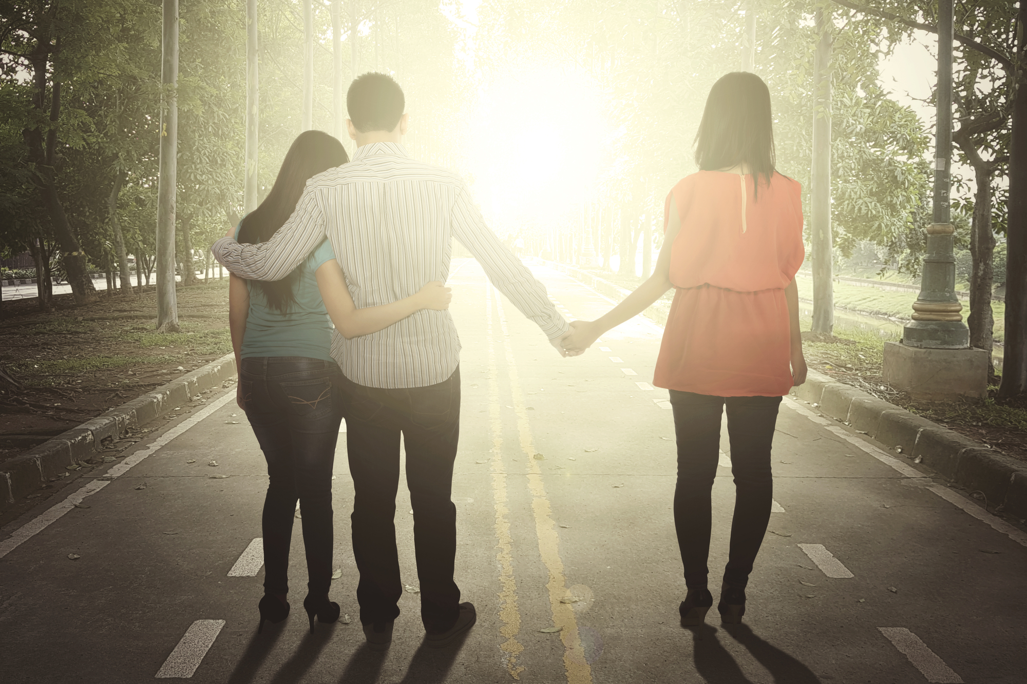 Three people walking down a sunlit road with trees on either side. A man in the middle has his arms around two women, one on his left wearing a teal shirt and the other on his right wearing a pink shirt. They hold hands and face away from the camera.