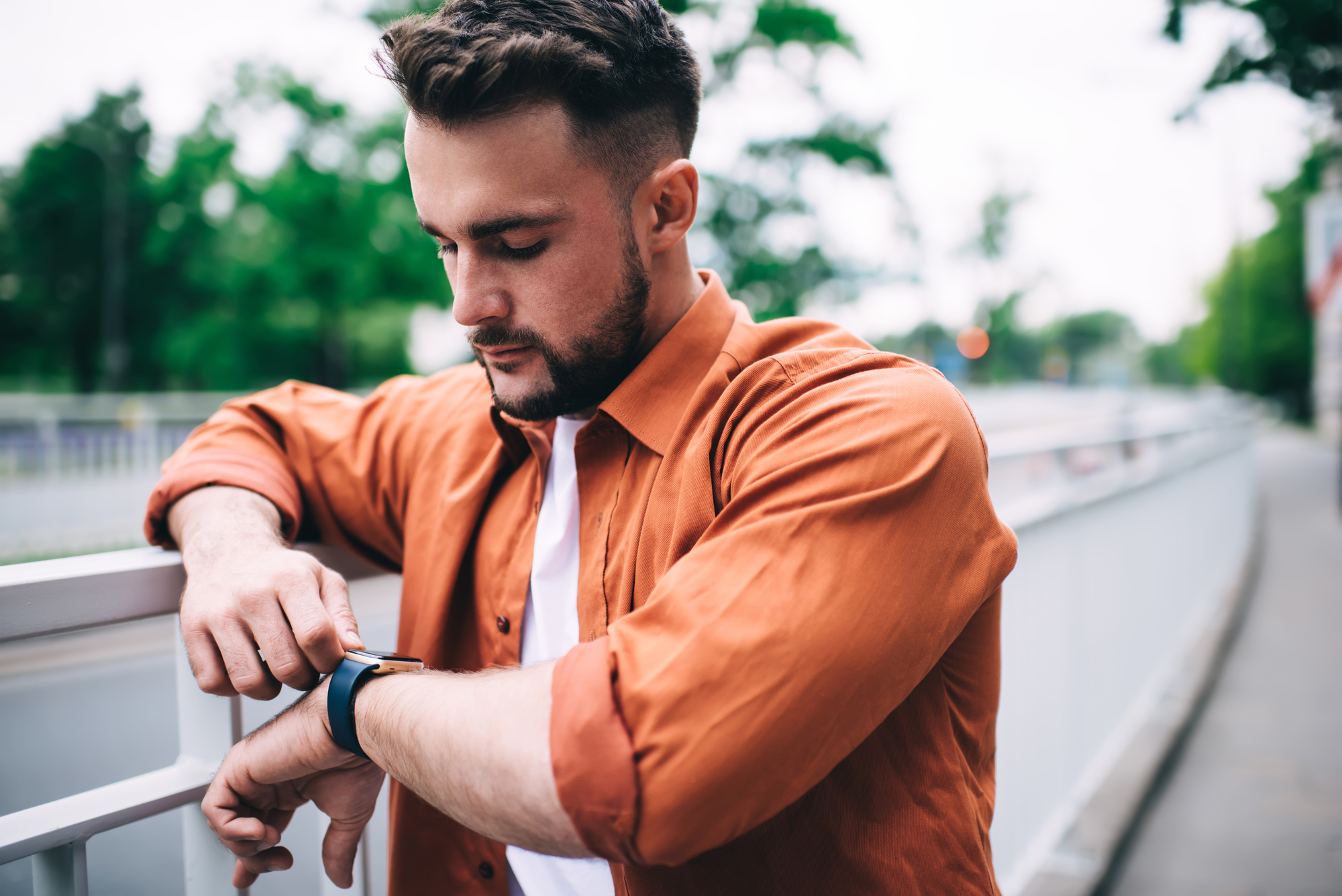 A man with short hair and a beard, dressed in a white t-shirt and an orange button-up shirt, stands outdoors by a railing, looking at his smartwatch. Trees and a pedestrian path are visible in the background.