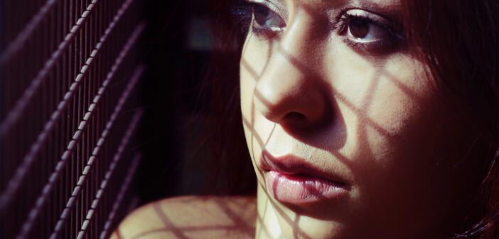 A woman with long hair looks out pensively, with shadows from a grid pattern cast across her face. She appears thoughtful, and the light highlights the intricate shadows, creating a dramatic effect on her expression.