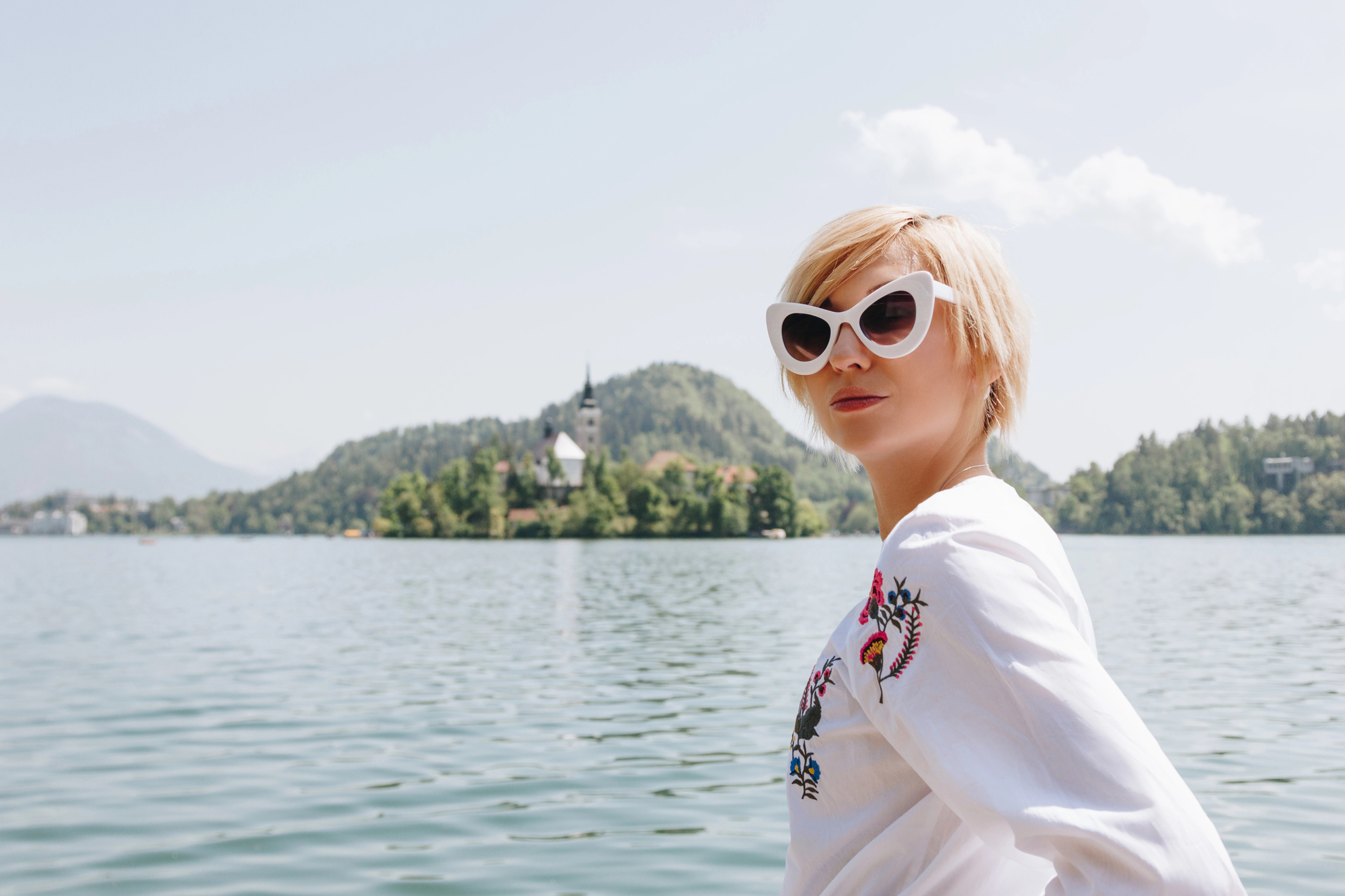 A woman with short blonde hair and wearing white cat-eye sunglasses stands in front of a serene lake. She wears a white blouse with colorful embroidery as she looks over her shoulder. In the background, there's a church on an island and hilly landscape.