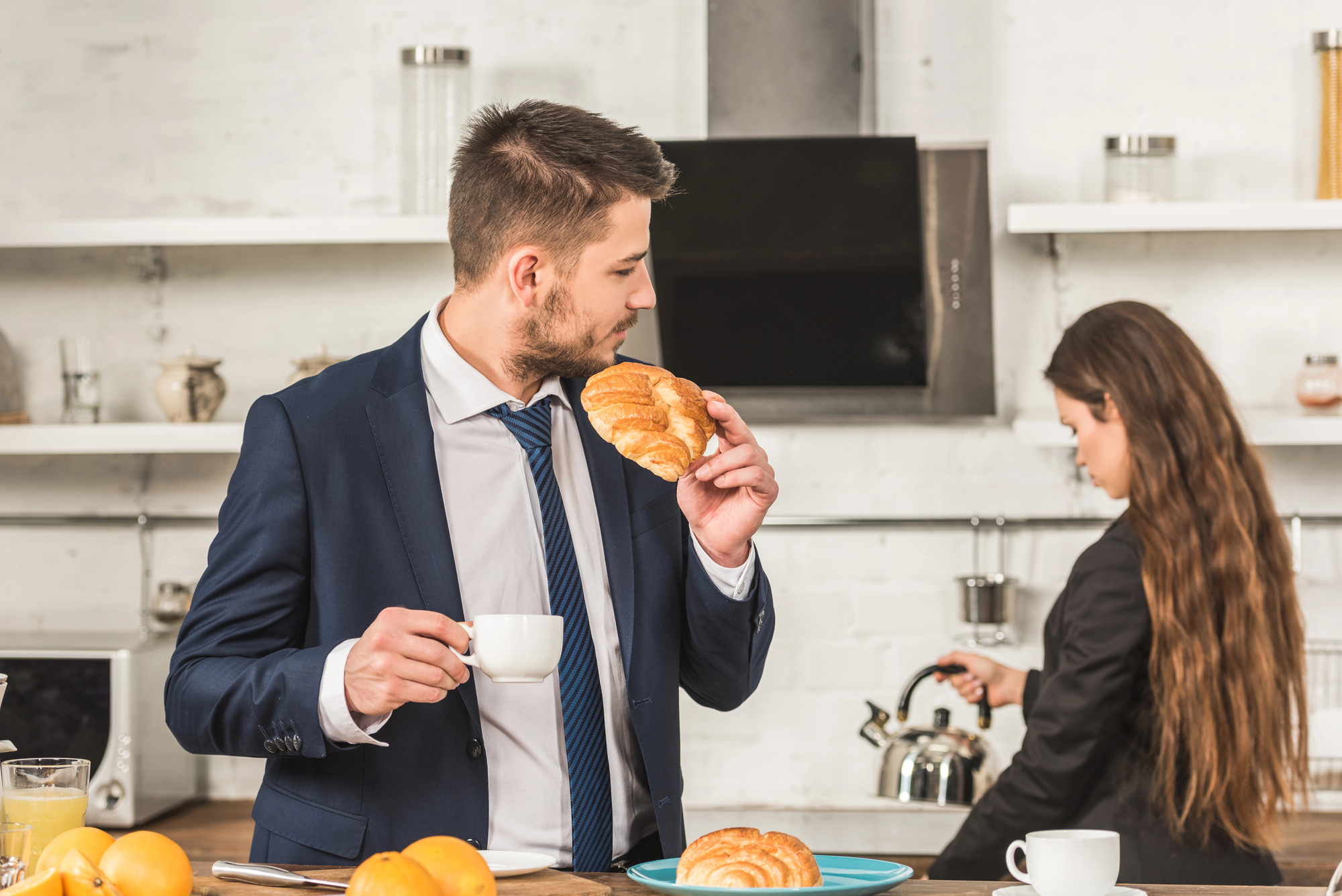 A man in a suit enjoys a croissant and holds a cup of coffee while standing in a modern kitchen. A woman in the background, also dressed in business attire, pours herself a drink from a kettle. Croissants and a plate of fruits are on the counter.
