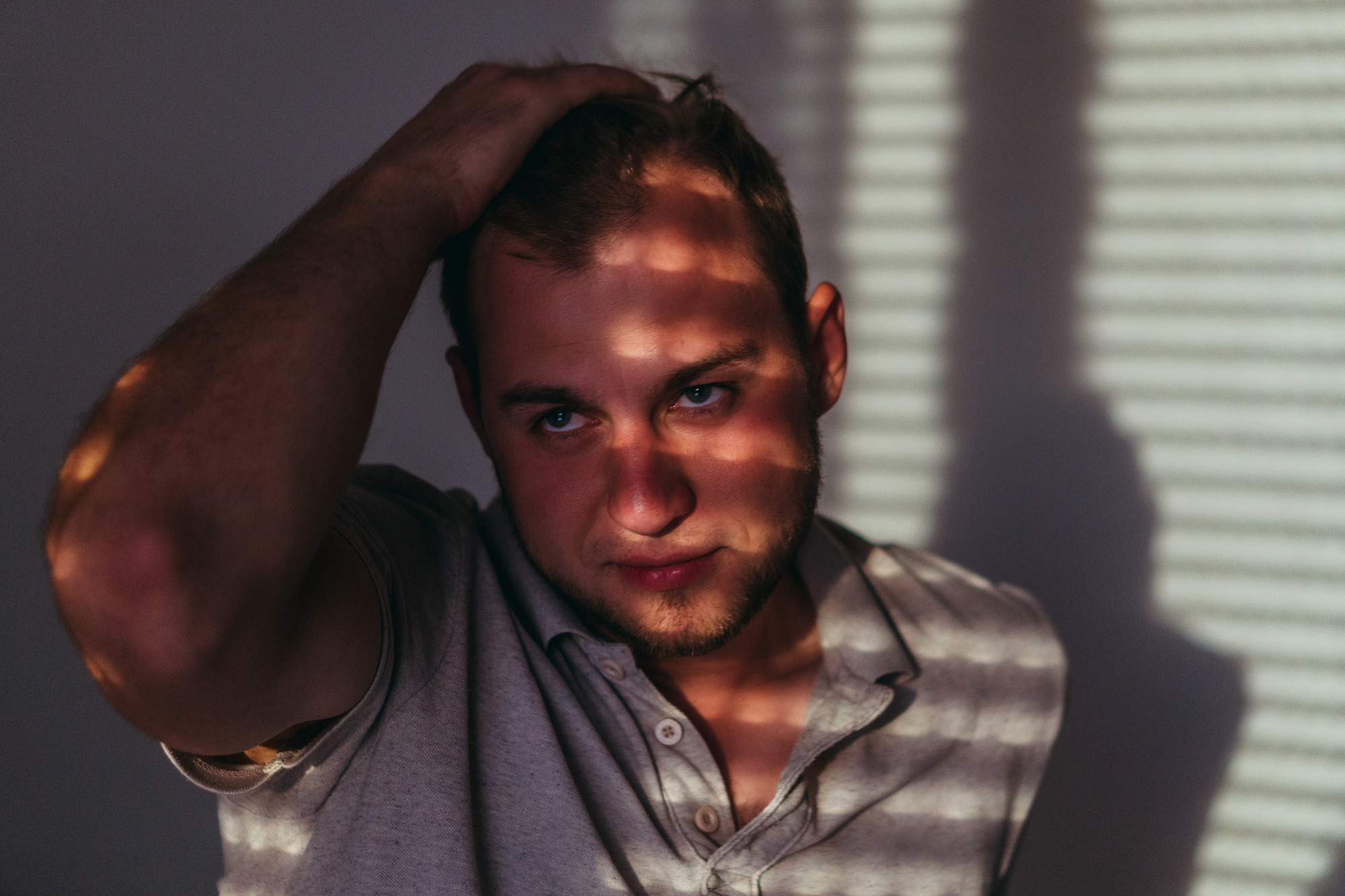 A man with short hair stands in partial shadow, touching his head as sunlight filters through blinds, casting striped patterns on his face and shirt. He wears a light-colored polo shirt and gazes slightly downward with a contemplative expression.